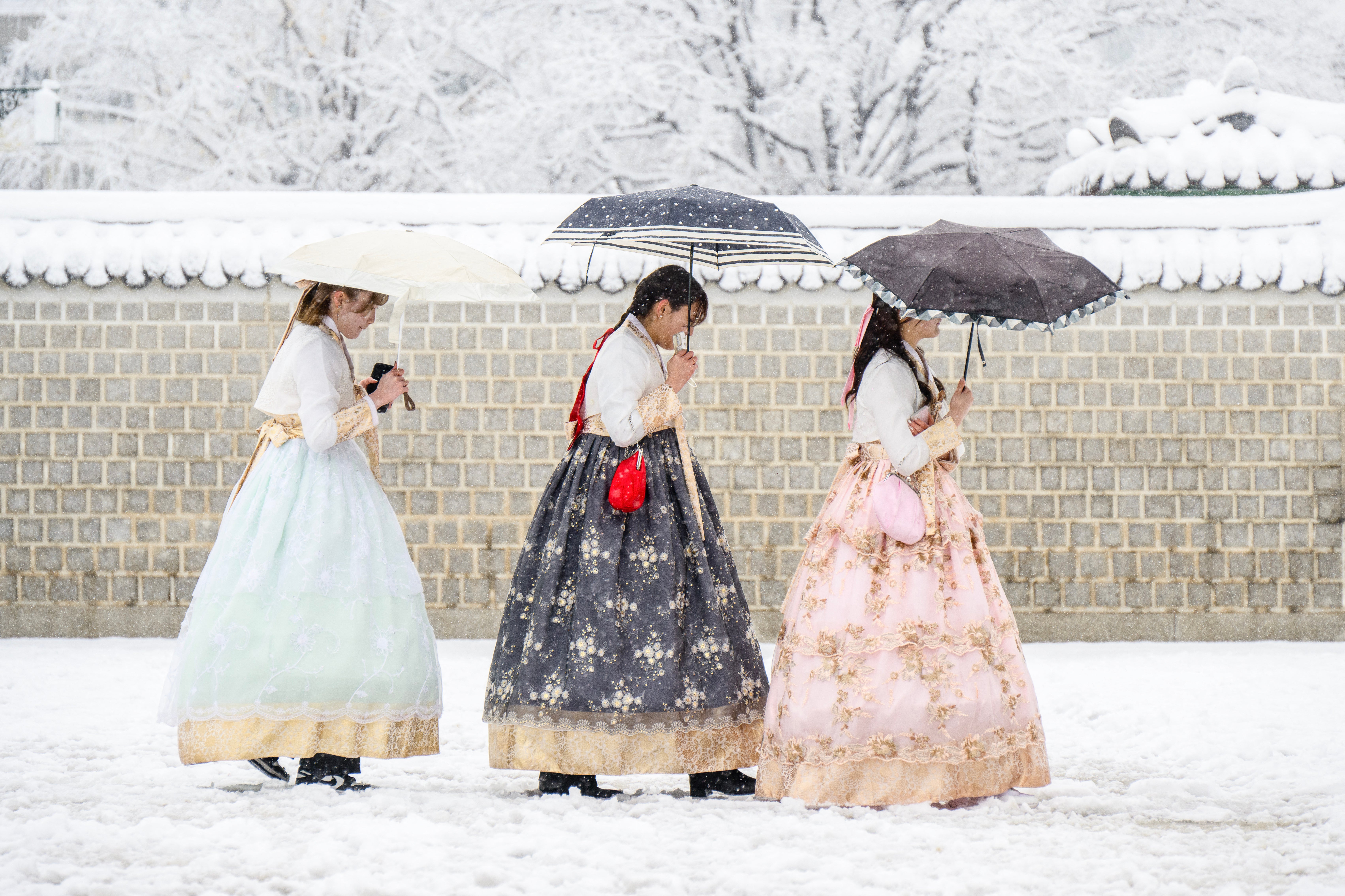 Individuals wearing traditional hanbok dresses trudge through the snowy conditions