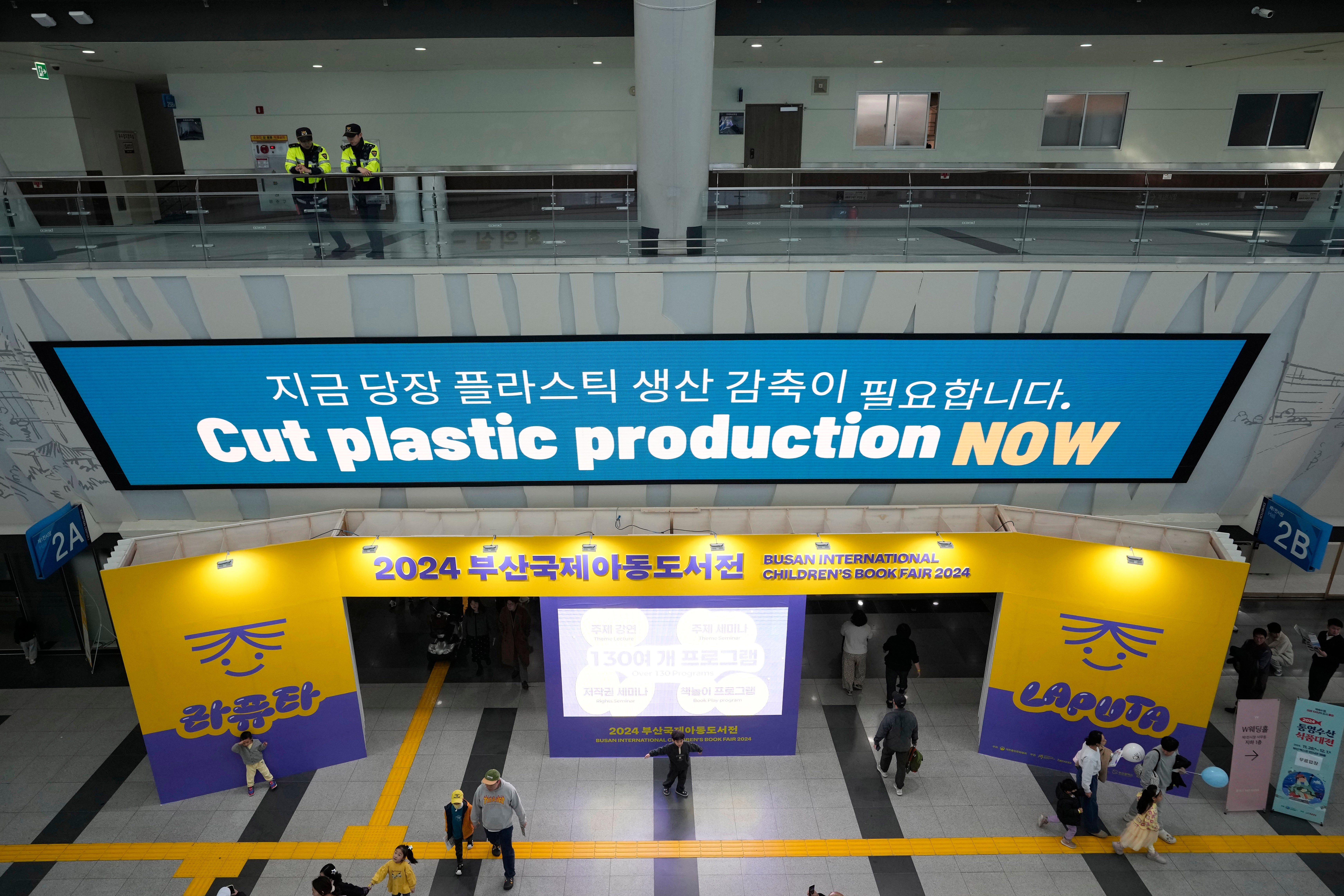 <p>People pass under an display board calling for a reduction in plastic production near the venue for the fifth session of the Intergovernmental Negotiating Committee on Plastic Pollution in Busan, South Korea</p>