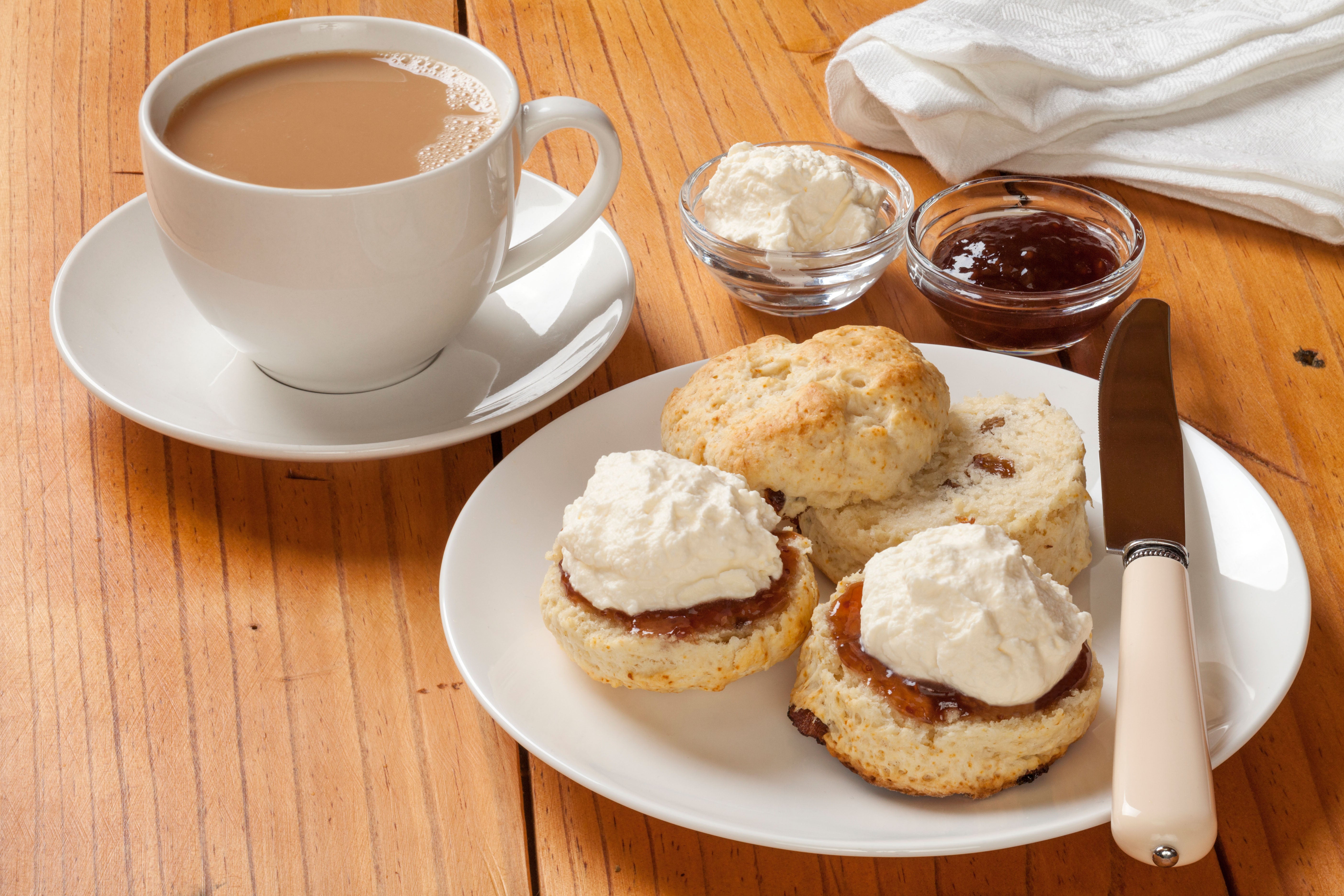 A plate of three scones with jam and cream next to a cup of tea with a saucer