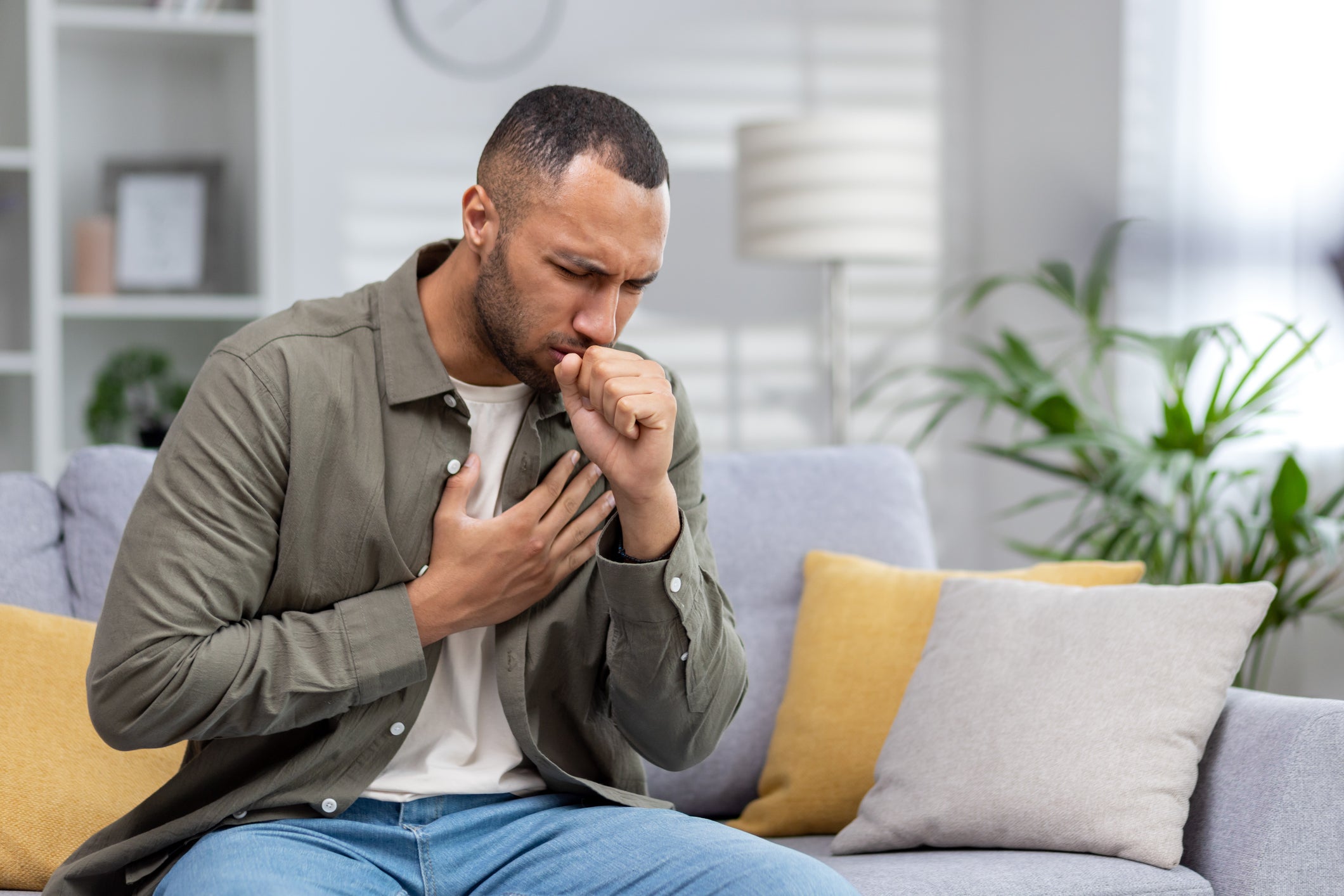 A young Black man coughs as he sits on a couch. Black people, women, and those between the ages of 18 and 60 are more likely to report not taking their asthma medications as prescribed because of the price tag