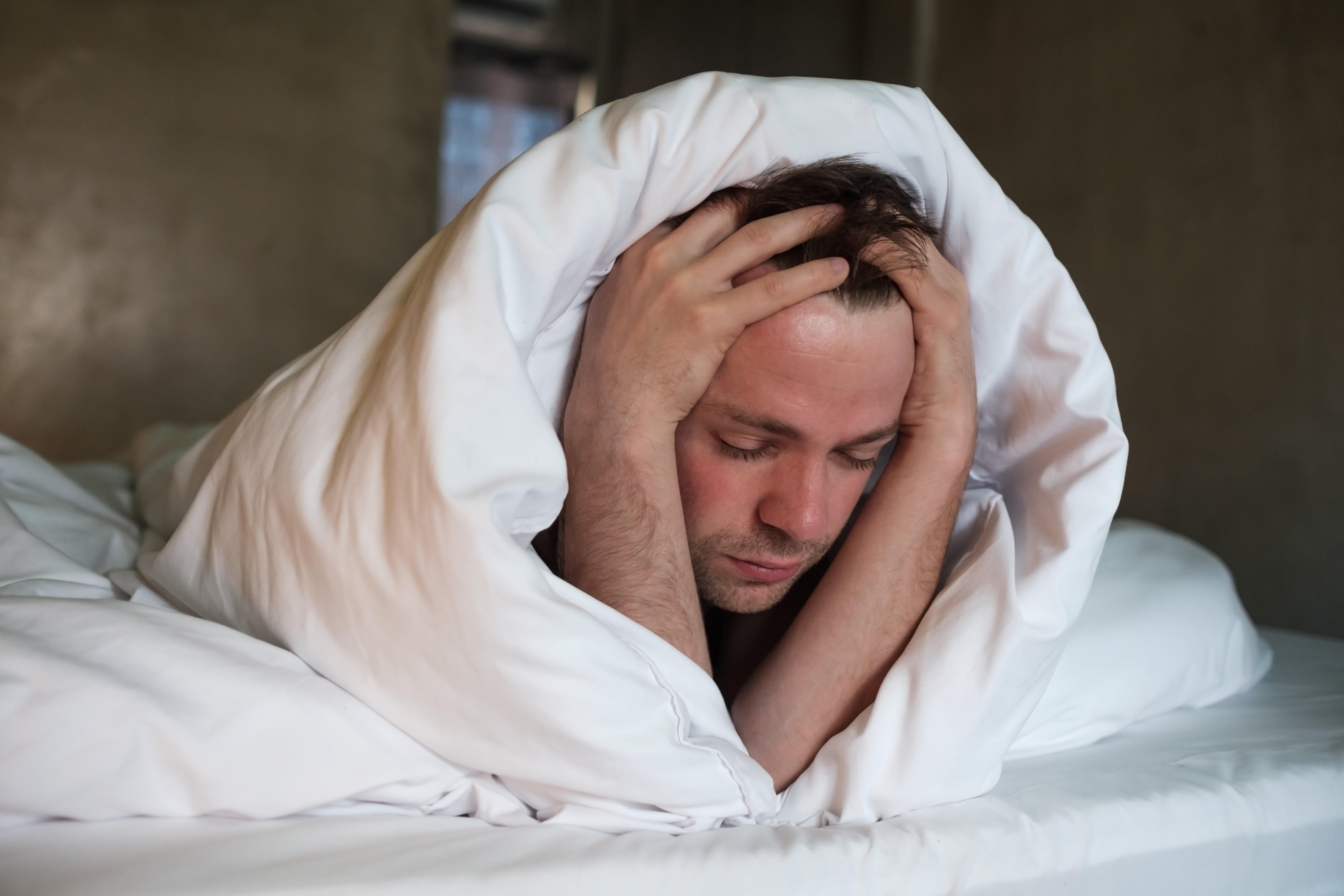 Depressed man lying in his bed wrapped in a duvet with his hands on his head