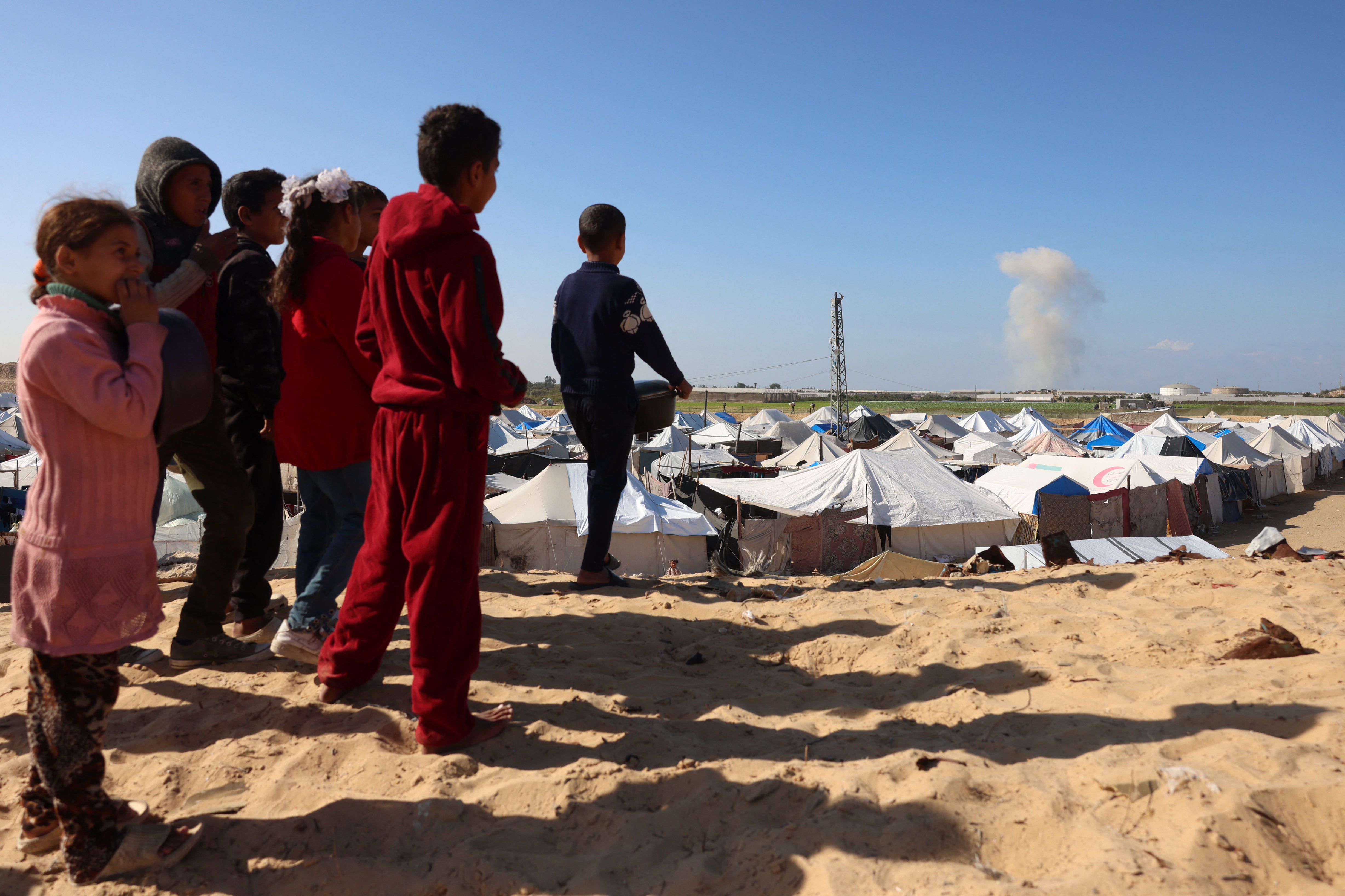 A group of Palestinian children watch smoke billowing as they wait for a food portion at a distribution center south of Khan Yunis in the southern Gaza Strip on December 17