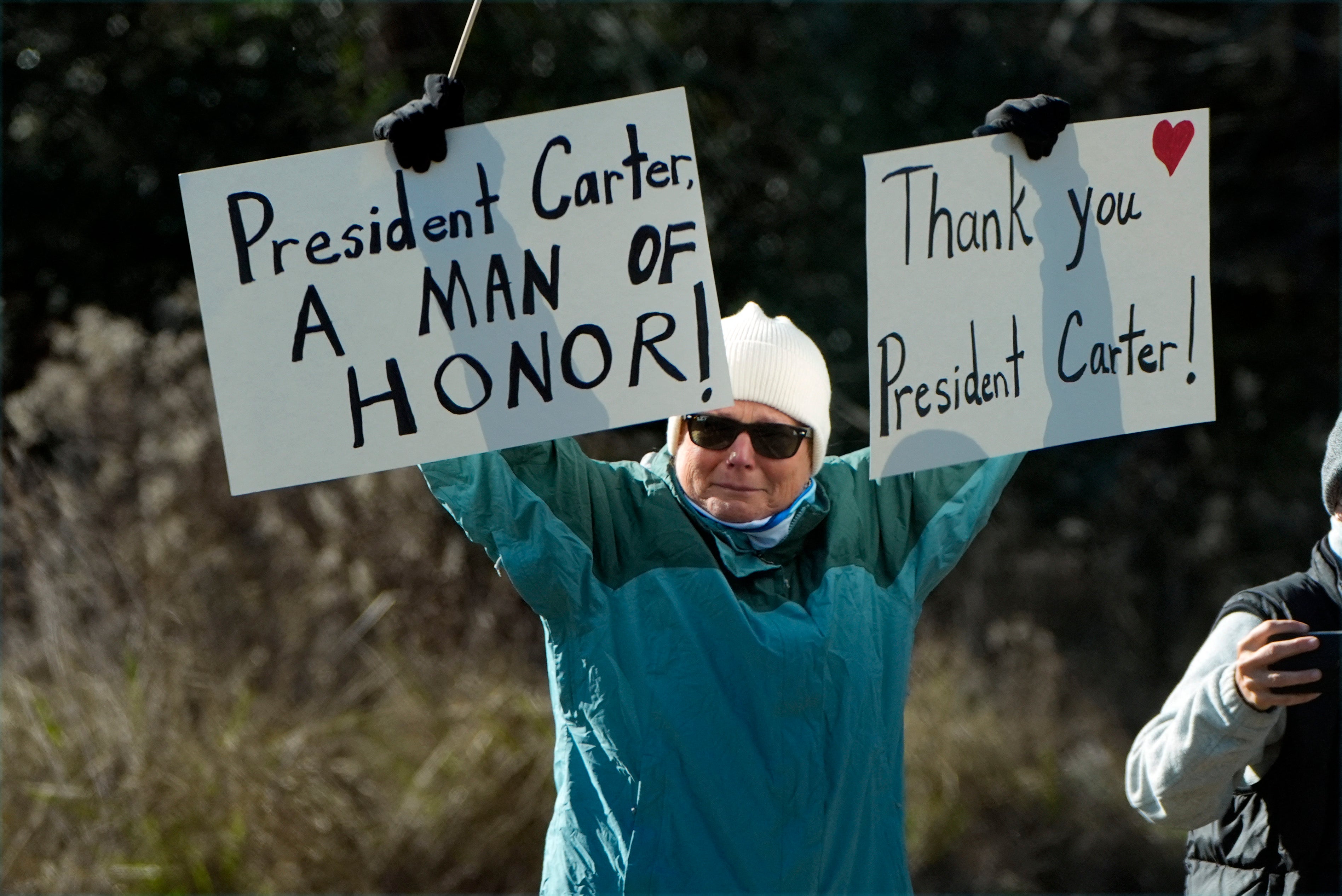 A mourner holds signs that read, ‘President Carter, a man of honor,’ and ‘Thank you President Carter,’ as the 39th president’s hearse drives through his hometown of Plains, Georgia