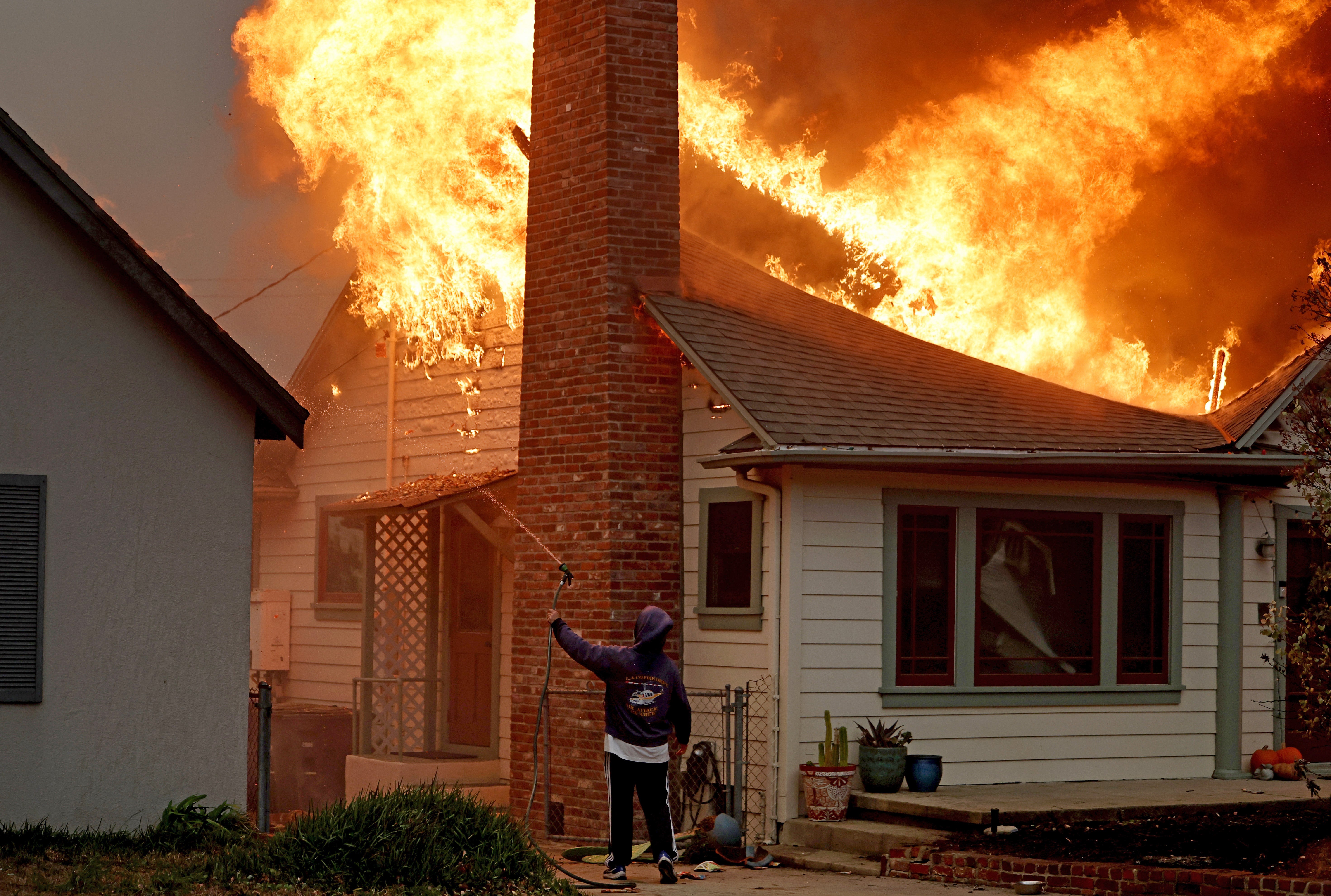 A person uses a garden hose in an effort to save a neighboring home from catching fire during the Eaton Fire on January 8 in Altadena, California