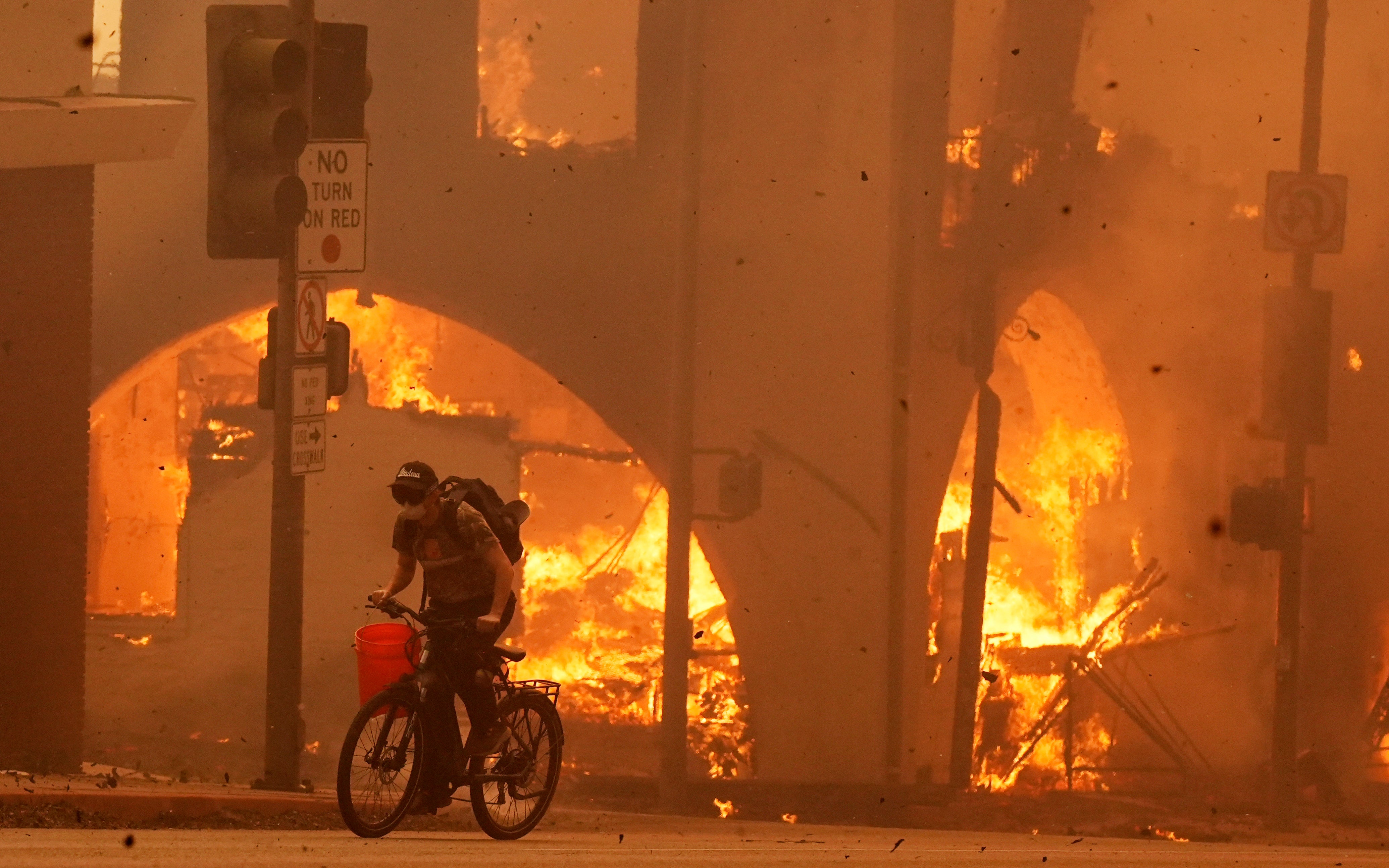 A cyclist pedals past a burning structure on Lake Avenue, Wednesday, Jan. 8, 2025, in the downtown Altadena section of Pasadena, California.