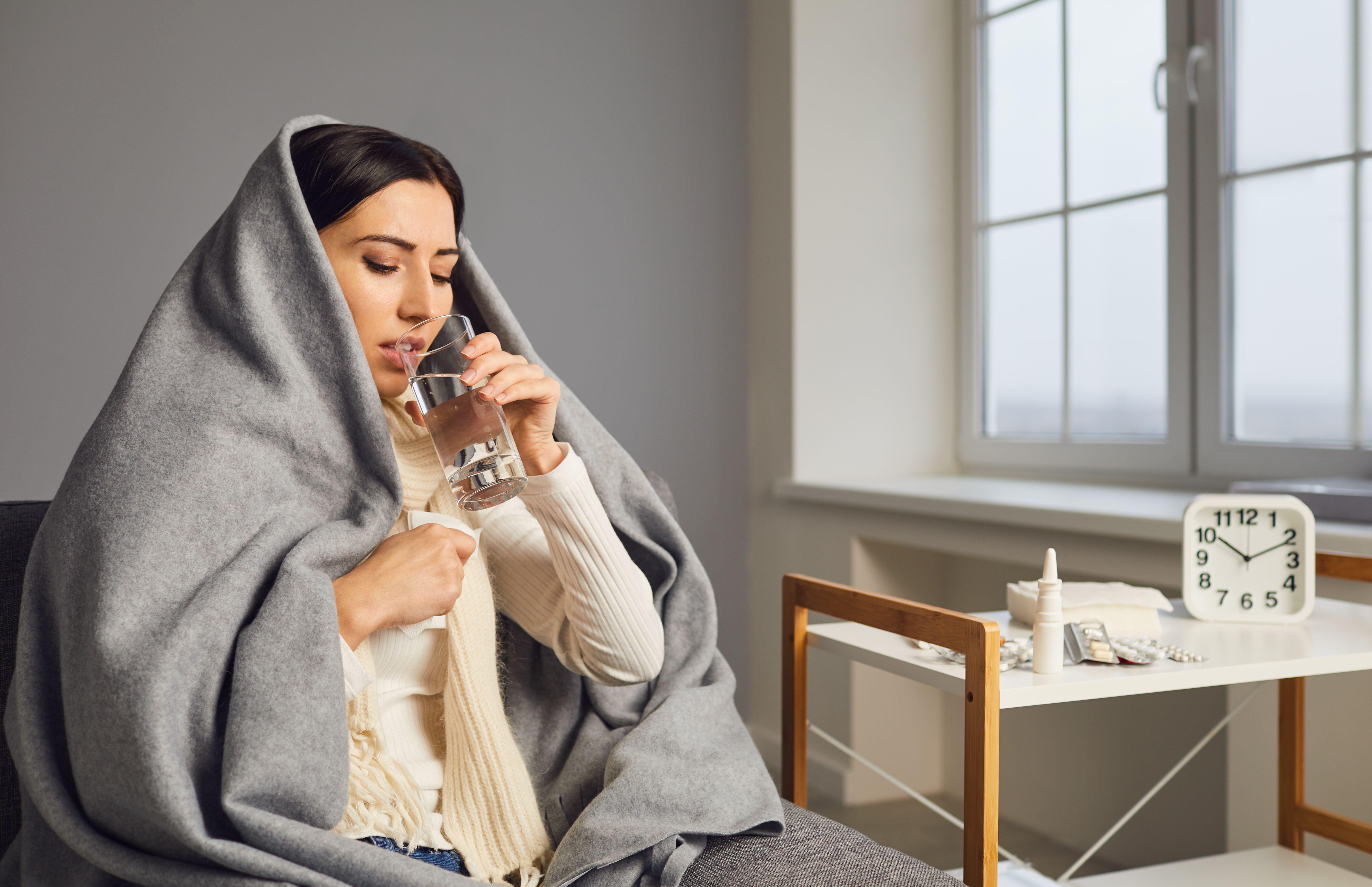 Side view of an ill young woman wrapped up in a blanket drinking water (Alamy/PA)