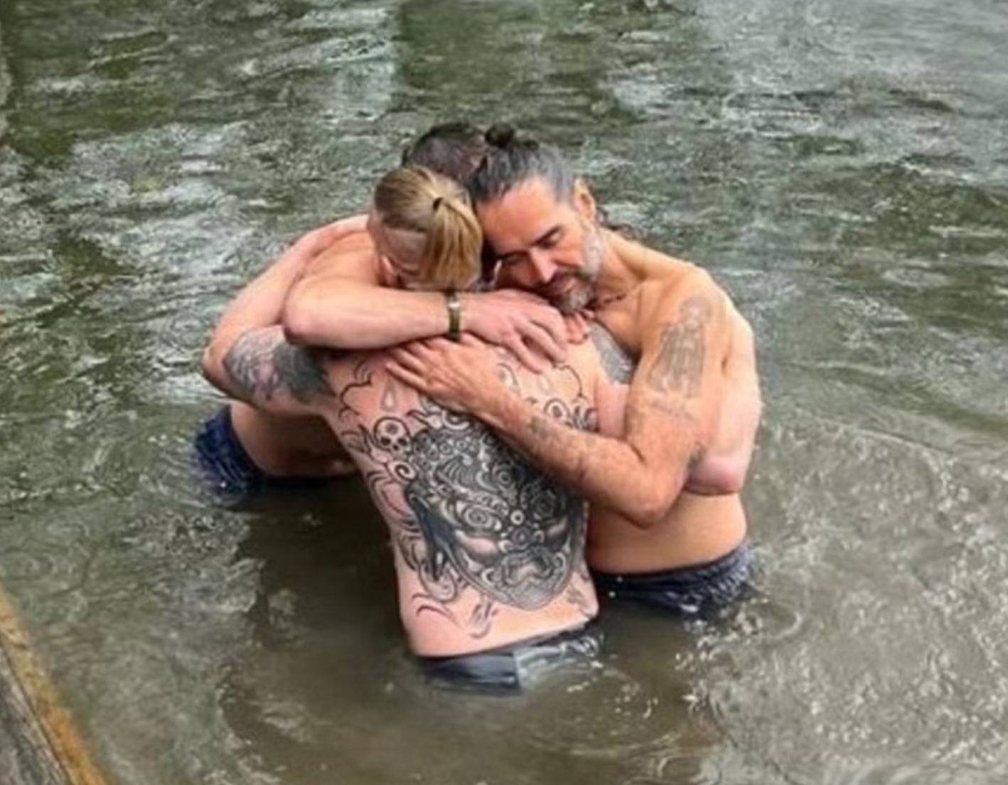 Bear Grylls and a third man hug Russell Brand after his baptism in the River Thames