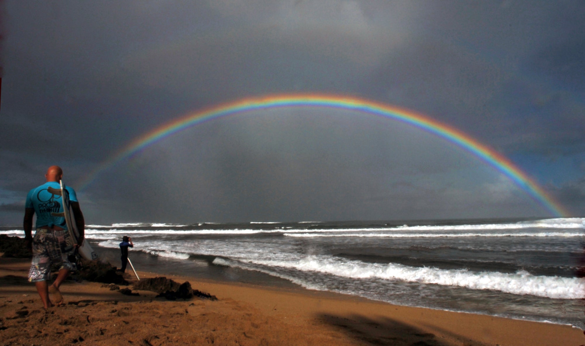 Professional surfers make their way to the water while a large rainbow crosses the ocean in Hawaii