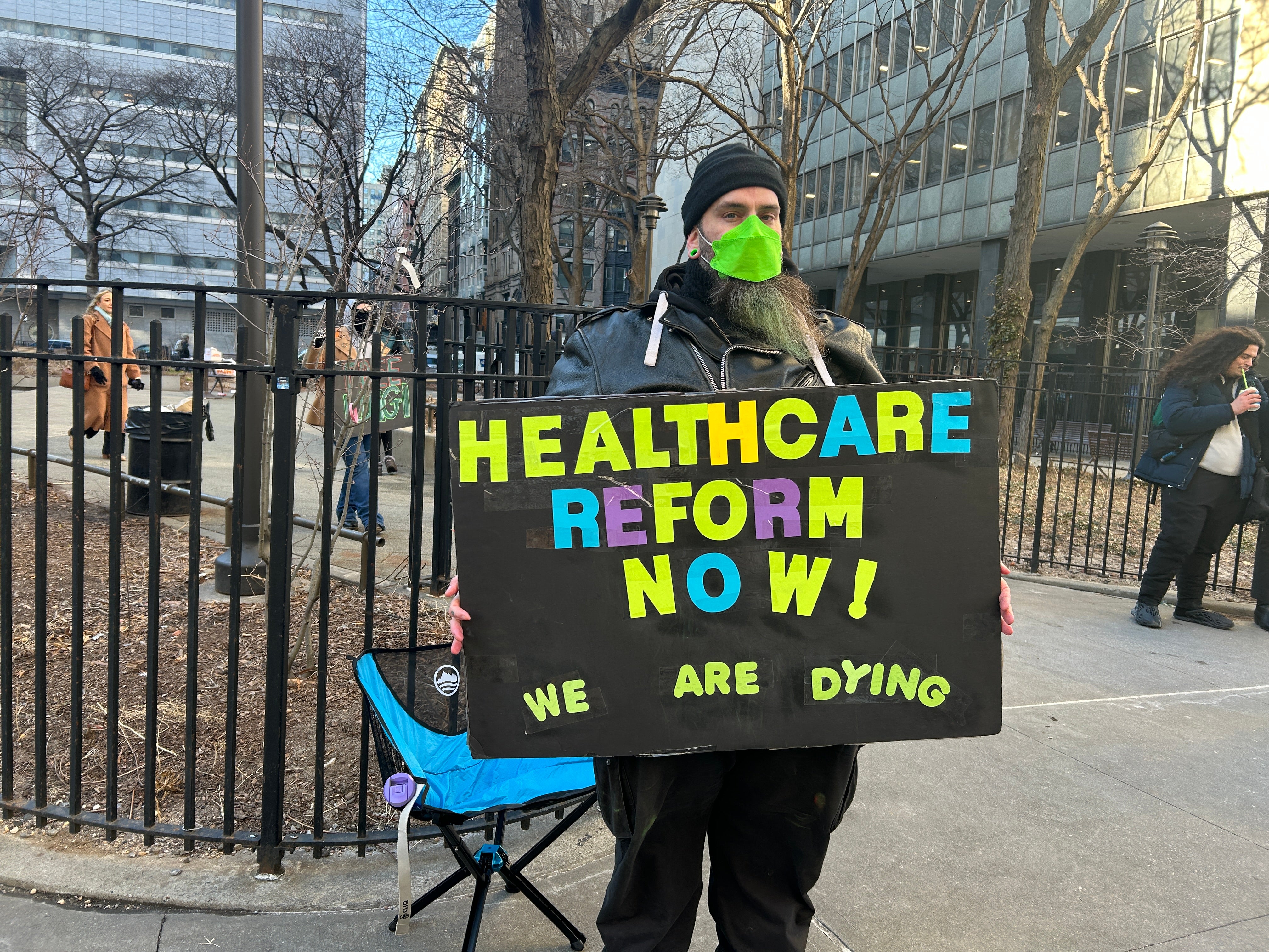 Church, wearing a green face mask, stands across from New York Criminal Court holding up a sign that reads: ‘Healthcare reform now! We are dying’