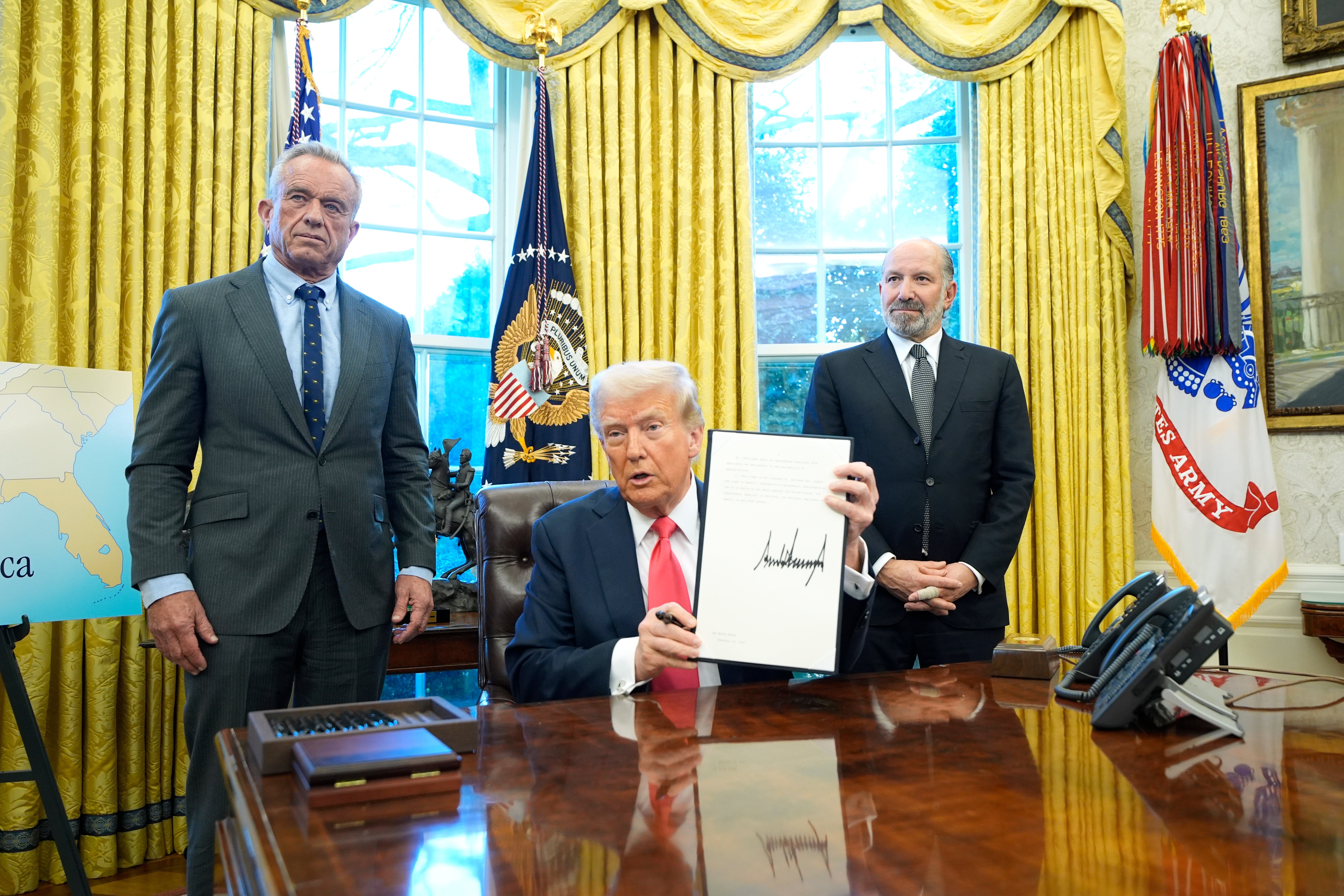 Robert F. Kennedy Jr., Donald Trump and Howard Lutnick stand in the Oval Office on Tuesday, where the president began advertising new merchandise