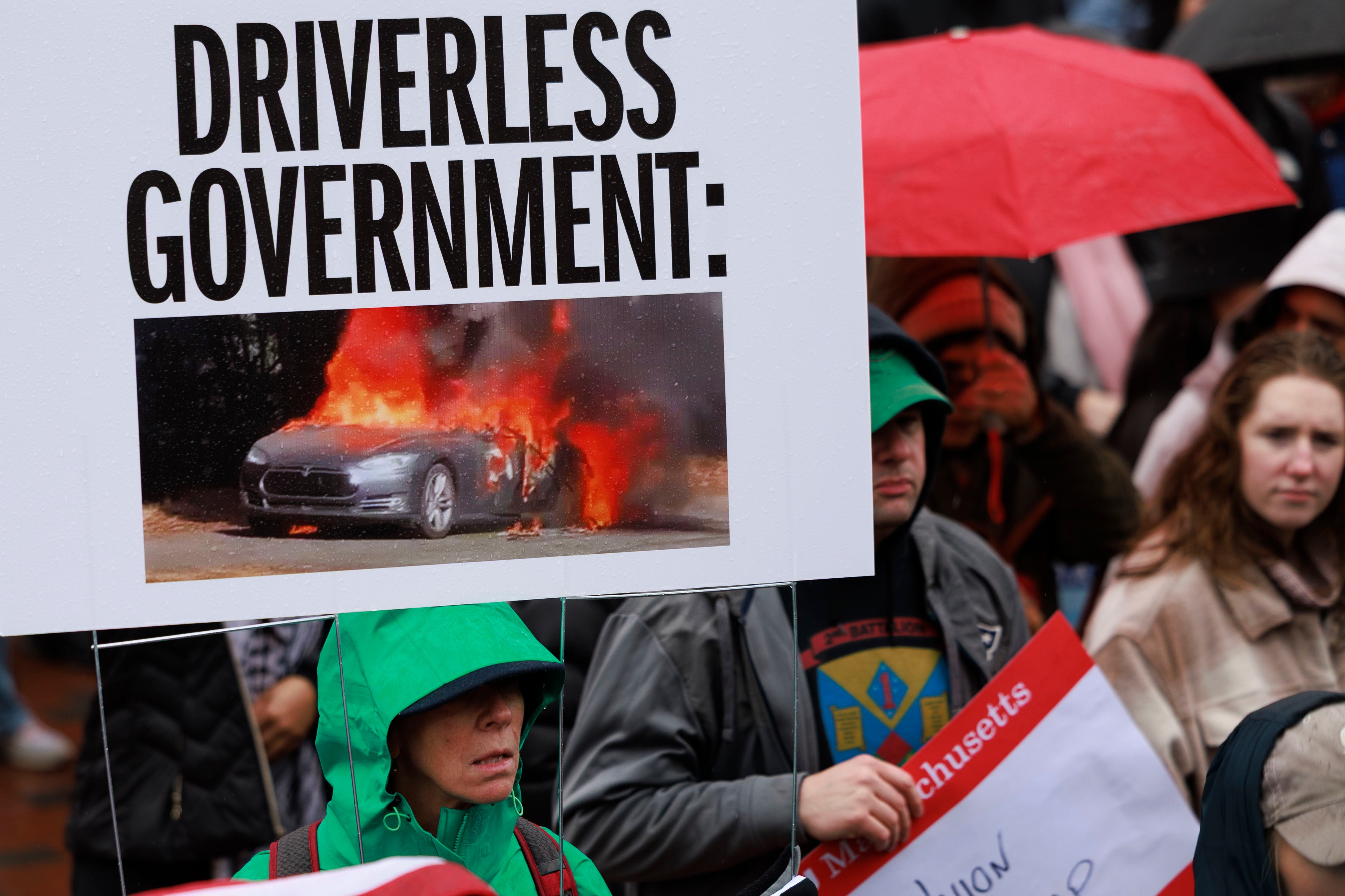 A woman carries a sign that reads 'Driverless Government,' showing a flame engulfed Tesla vehicle as she and thousands of people gather on Boston City Hall Plaza