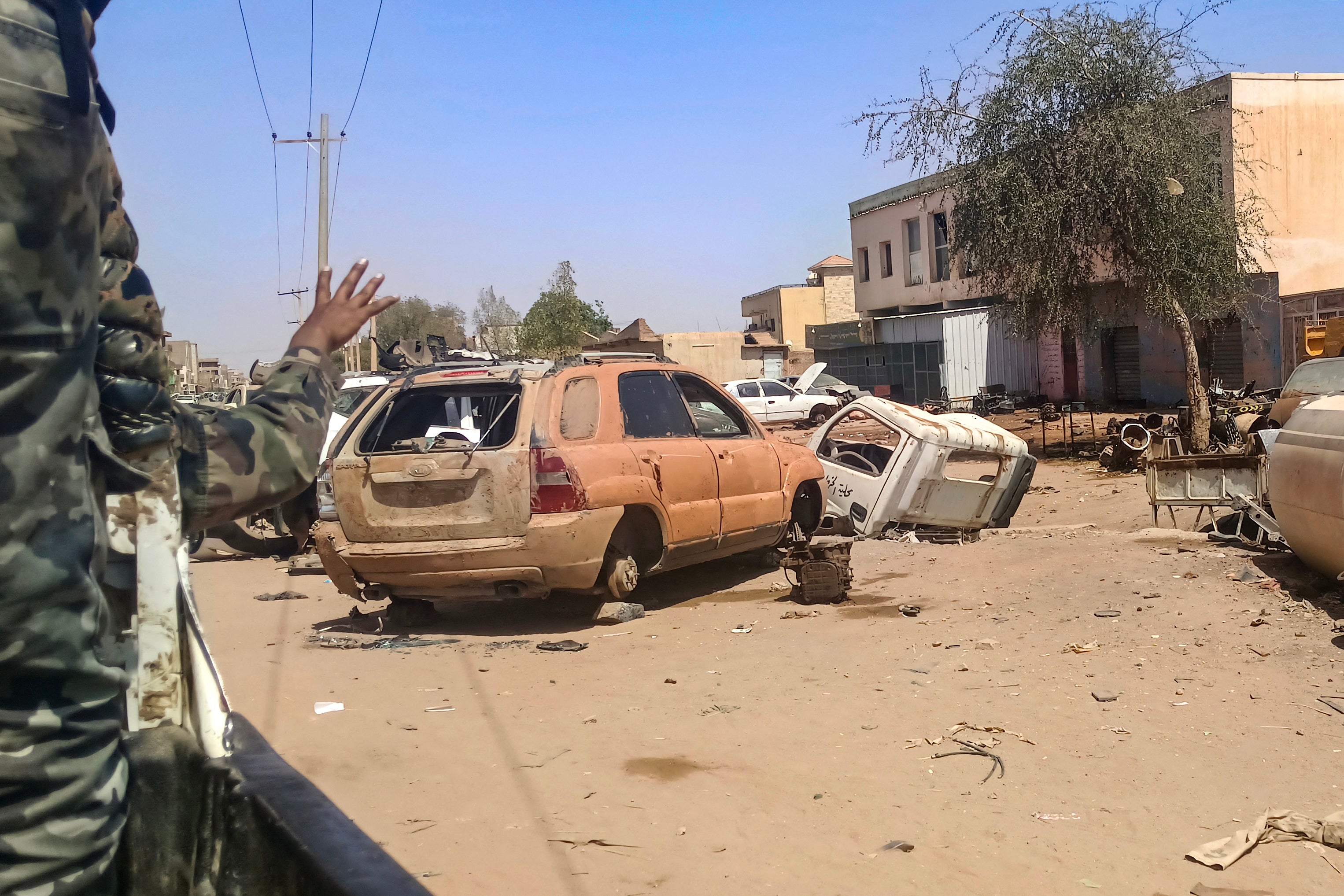 A Sudanese army soldier gestures from the back of a vehicle as it drives past damaged cars lying along a street in Khartoum