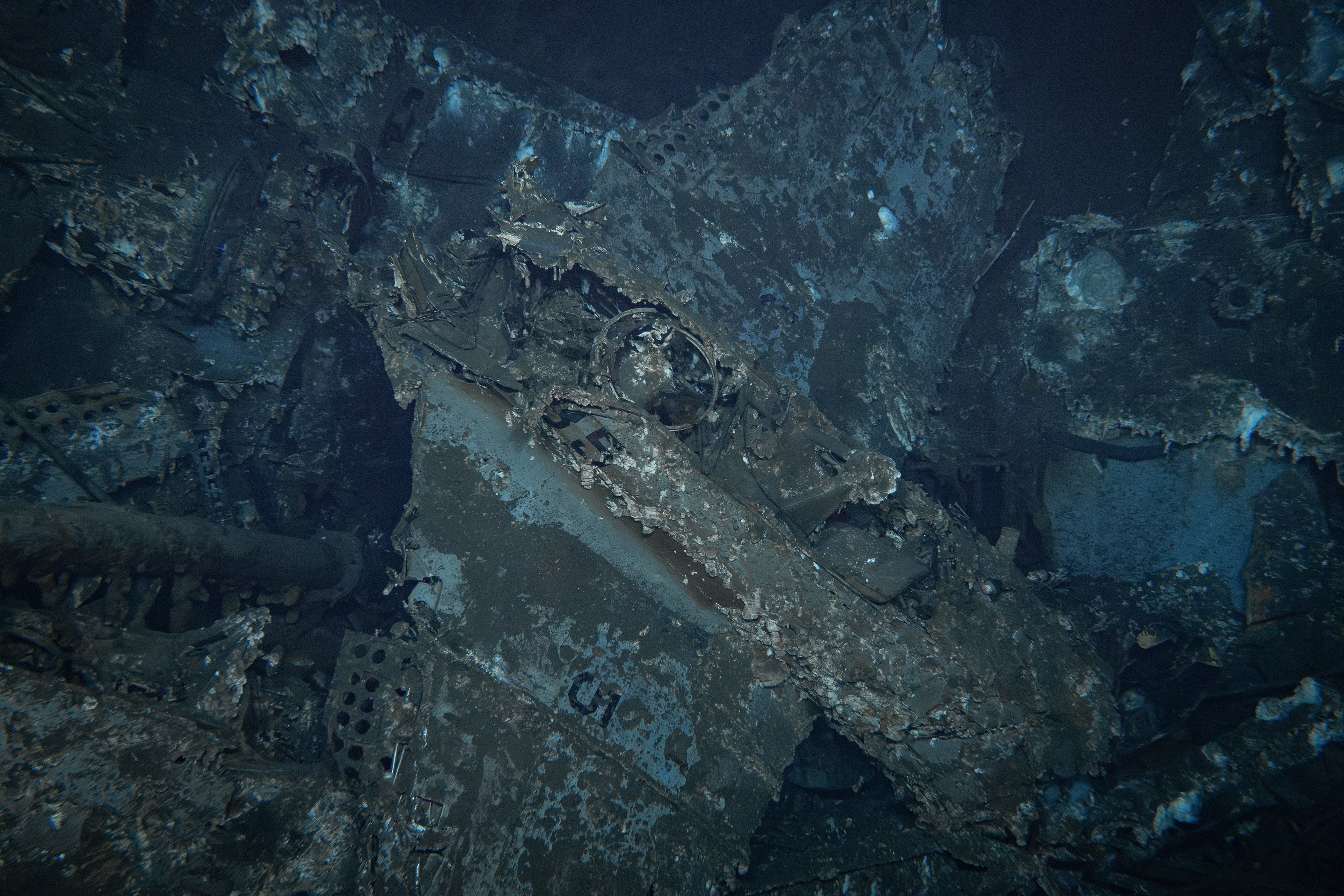 The battered wreckage of a Douglas SBD Dauntless dive bomber with legible markings on the fuselage beneath the gunner's ring mount (center of frame) and another "5" visible on the upper surface of the right wing (bottom). It is currently believed this aircraft is one of two battle-damaged planes from USS Enterprise's Bombing Squadron Six that landed aboard Yorktown following their successful attack on the Japanese carrier Kaga