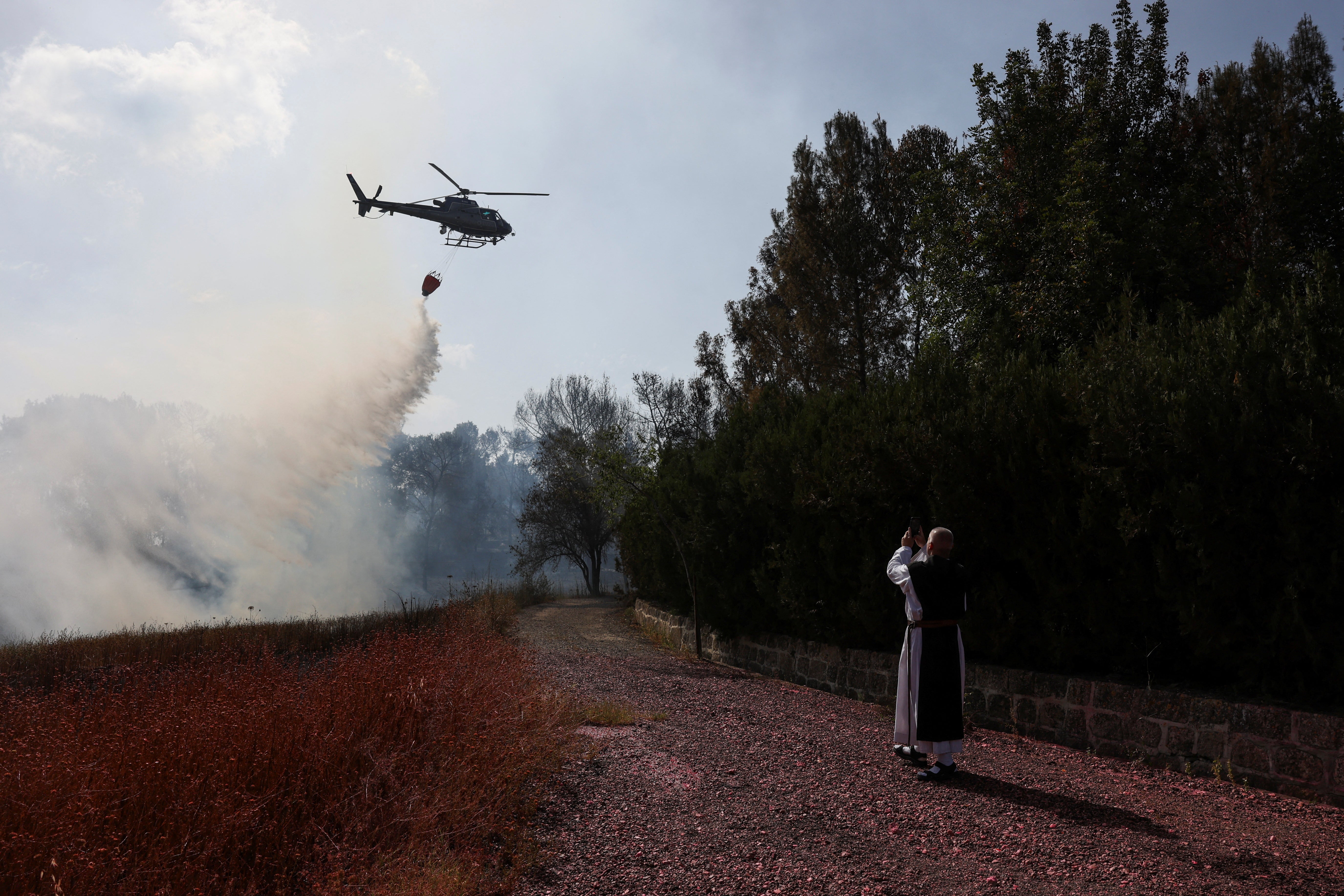 A Cistercian monk takes a picture of a helicopter dropping water near Latrun, a day after wildfires broke out due to extreme heat and winds, in central Israel on 1 May 2025