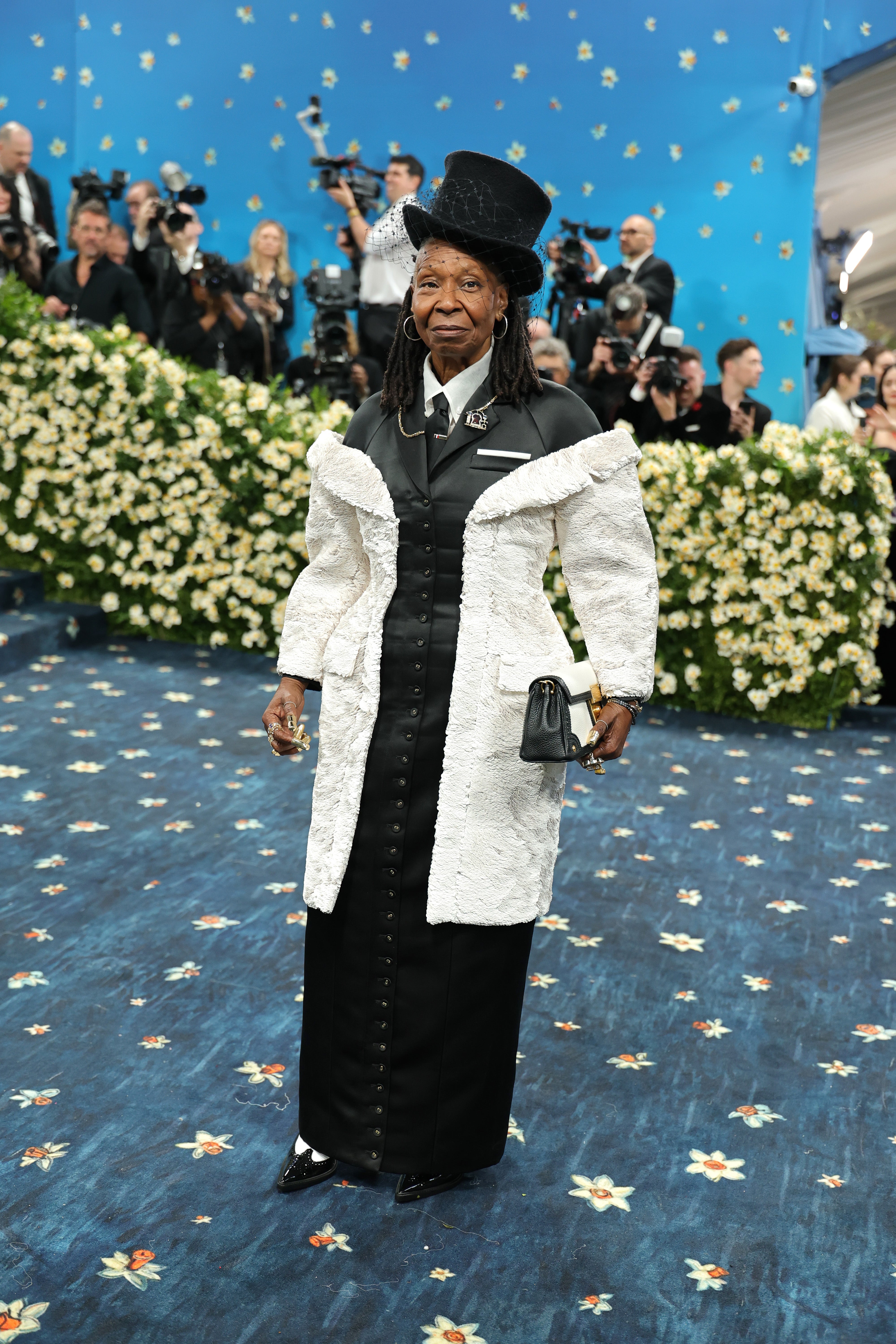 Whoopi Goldberg wore a black buttoned dress over a black tie and white shirt. Her look, created by Thom Browne, included a white, off-the-shoulder jacket, black top hat with a fascinator, and black and white purse