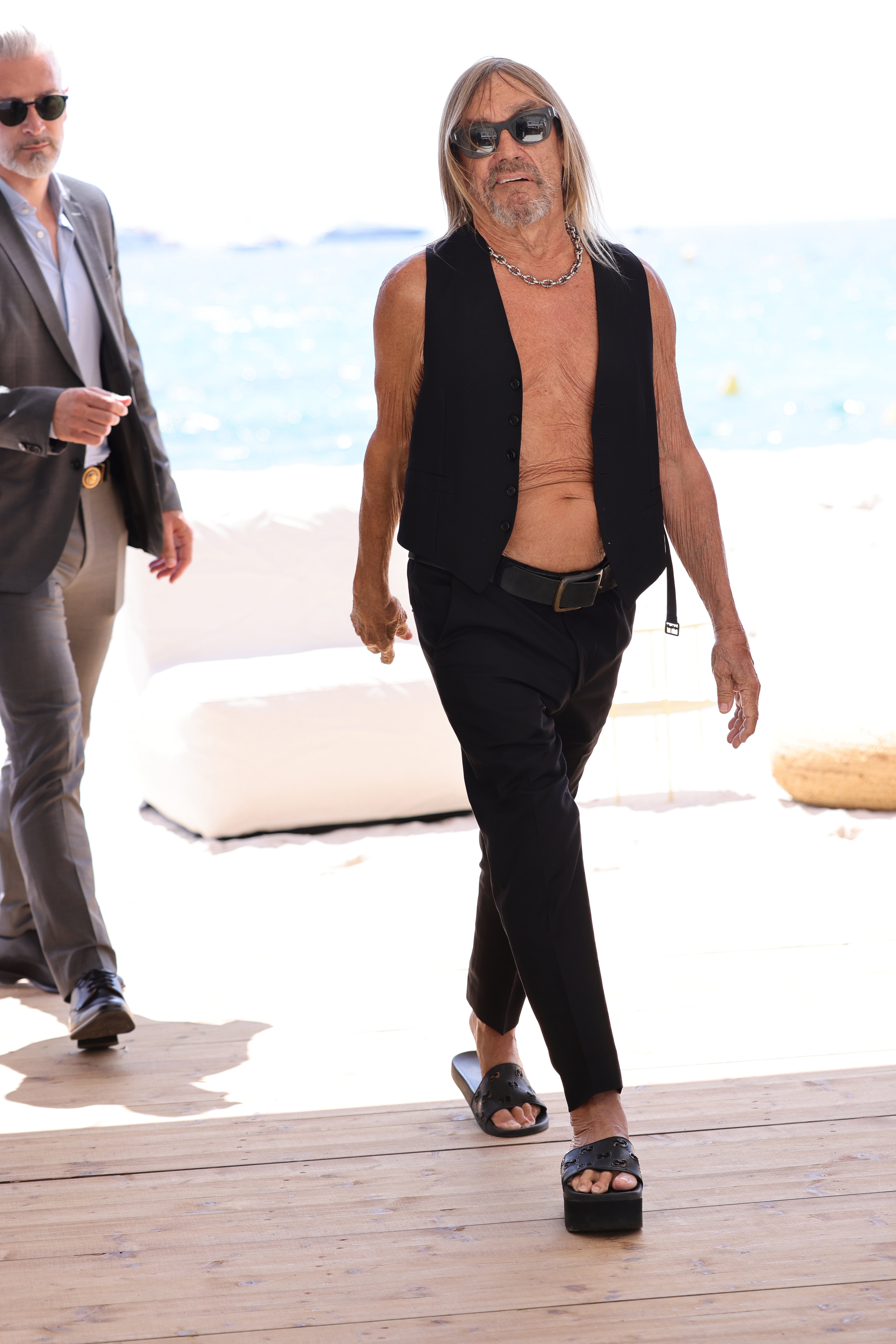 Singer Iggy Pop strutted along the beach in Cannes for the 2024 festival in a classic black vest, matching pants, sunglasses, and open-toe sandals