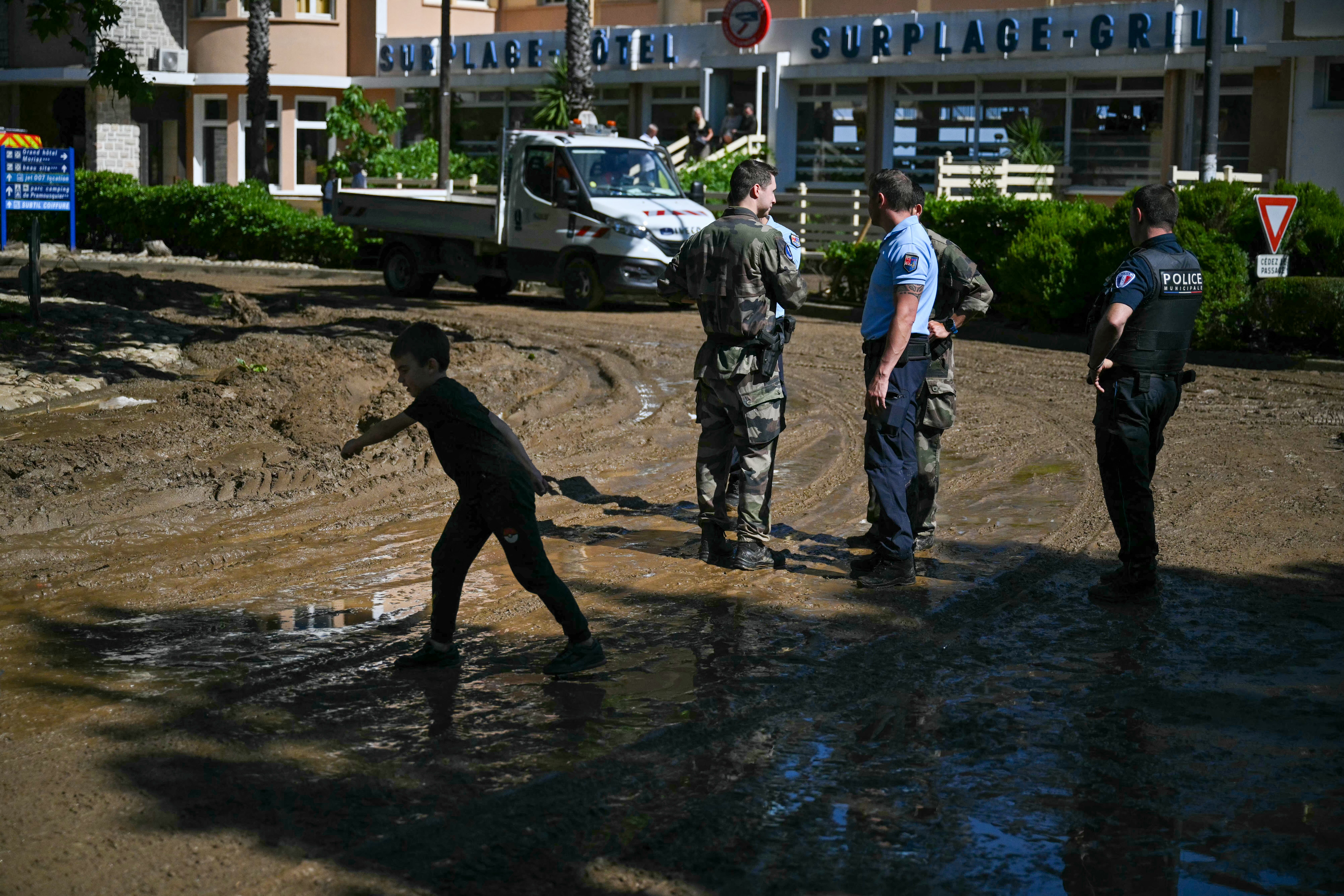 A child walks past French police officers on a mud-covered street following a flood caused by heavy rain in Cavaliere village