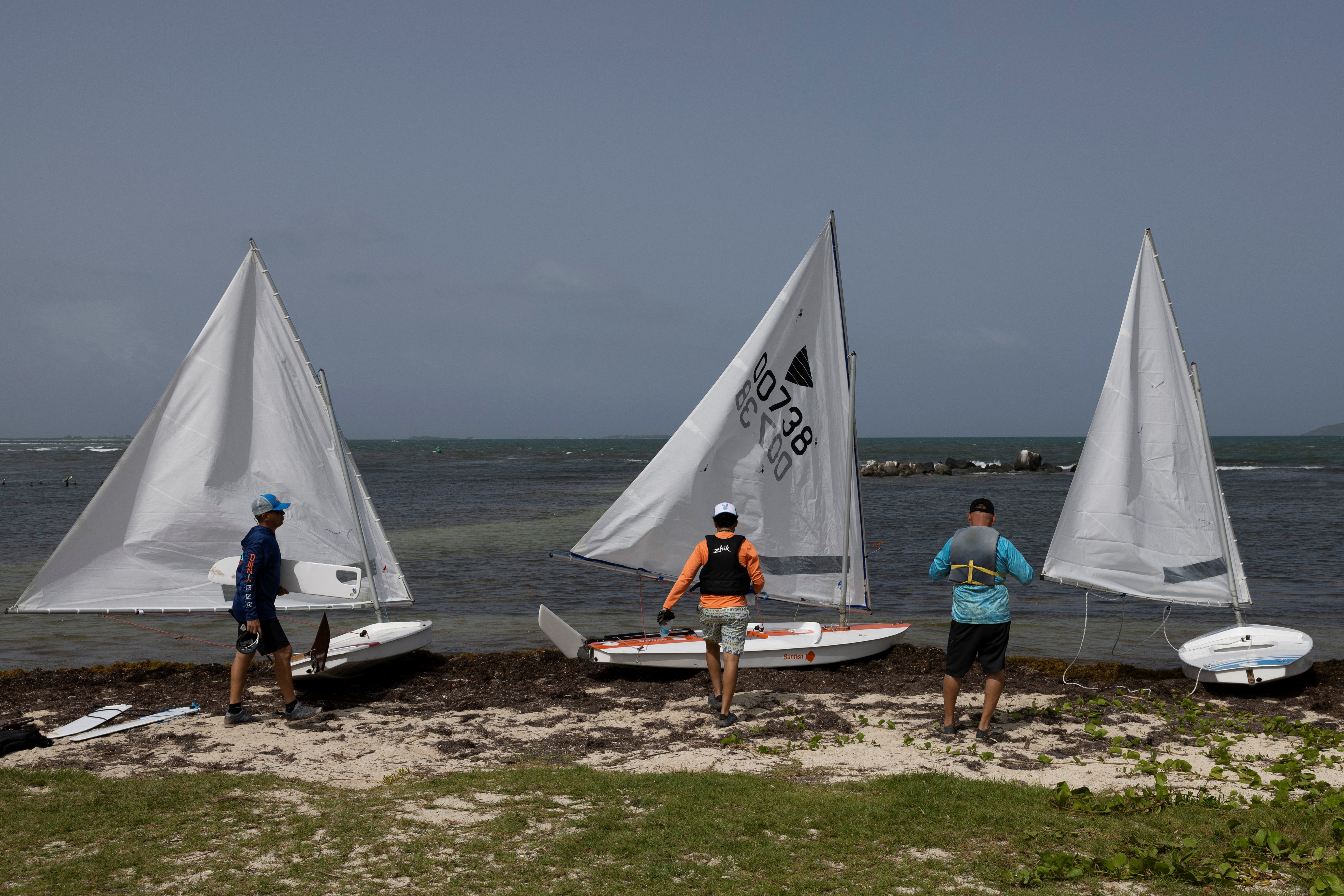 Men prepare to launch their sailboats on a beach covered in sargassum seaweed washed ashore