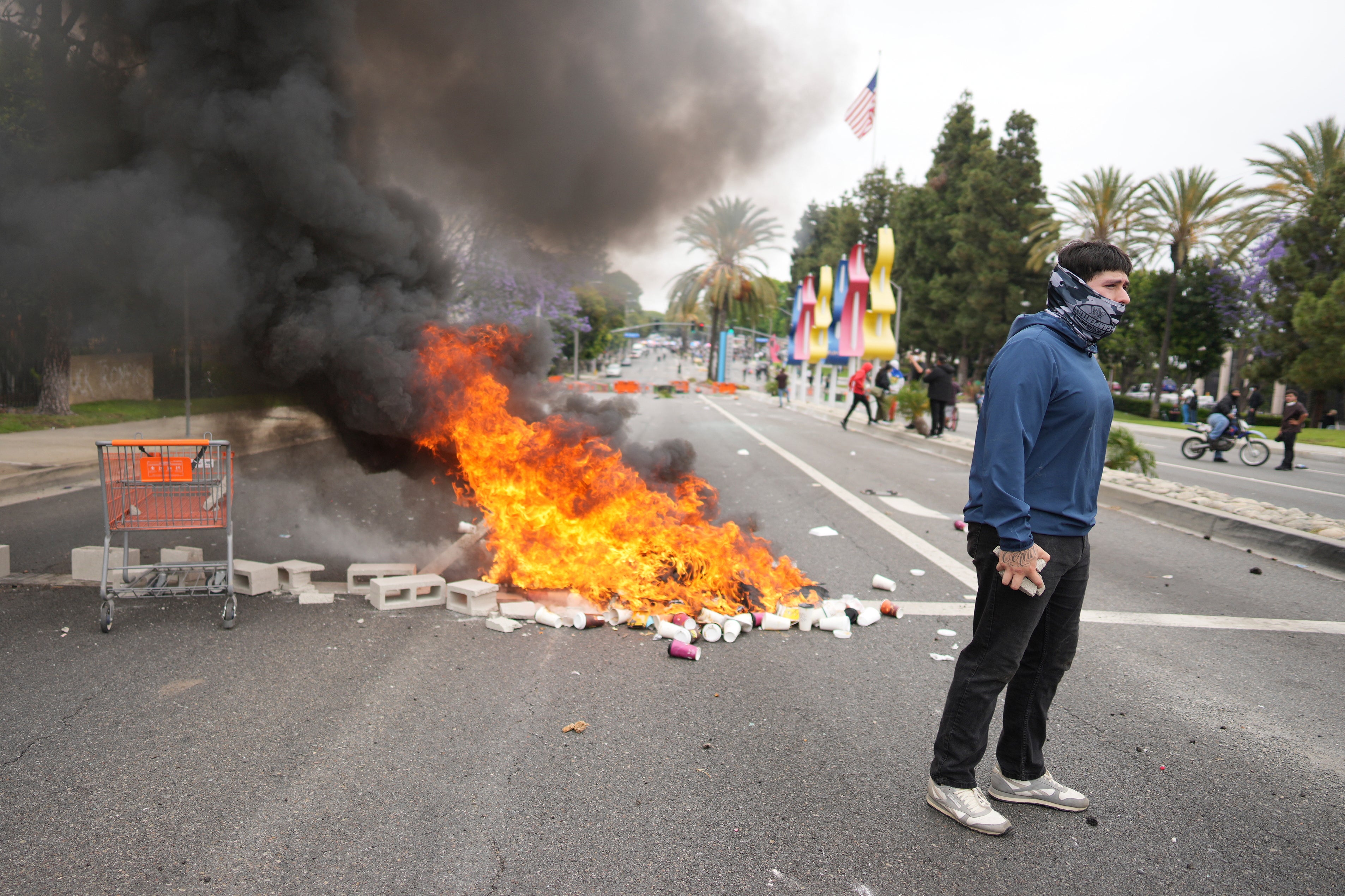 A fire burns as a protester confronts law enforcement personnel in riot gear and gas masks during a demonstration over dozens of people detained in an operation by ICE agents a day earlier in the Paramount area of Los Angeles