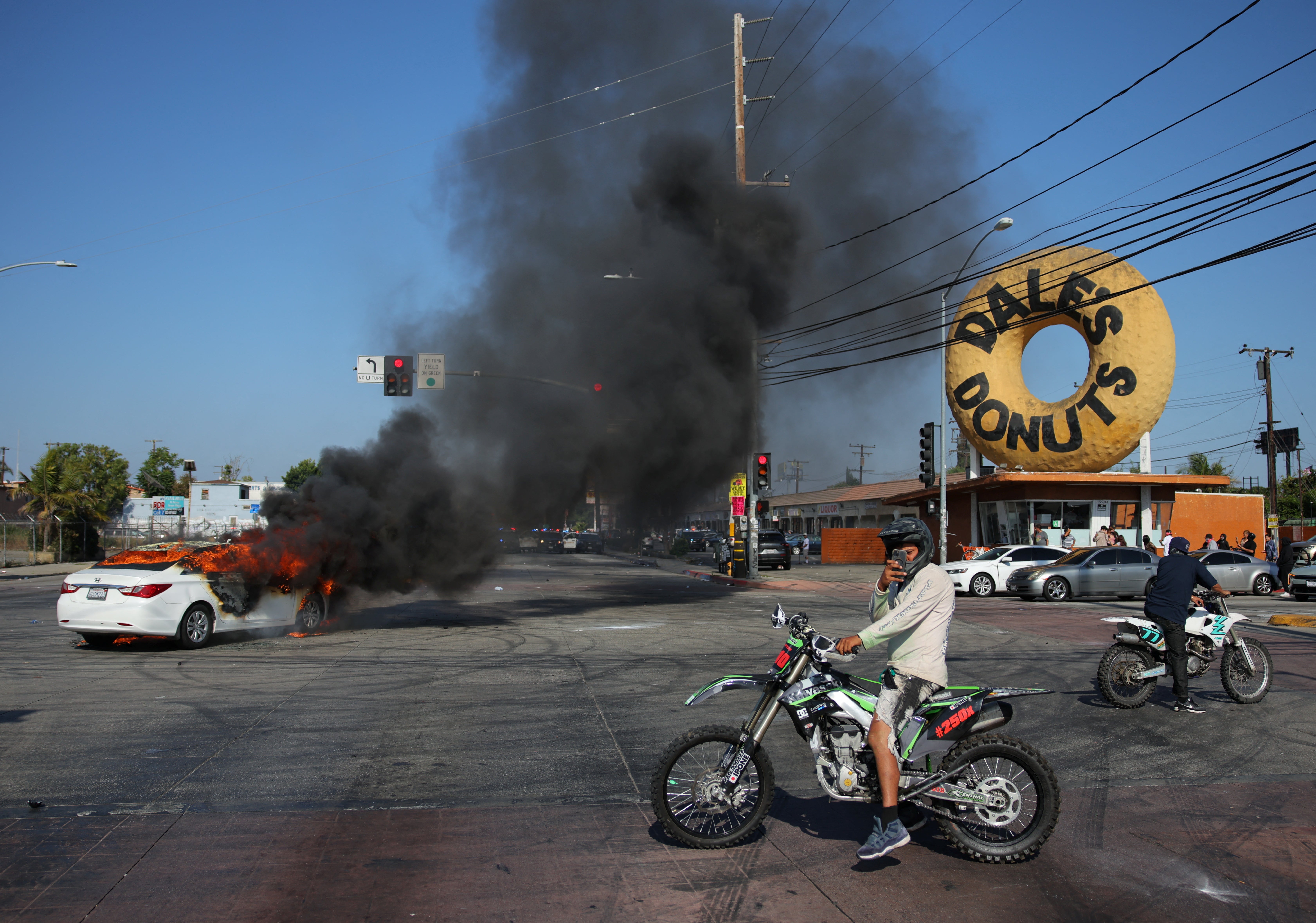 People ride motorbikes next to a car burning in flames during a standoff between police and protesters following multiple detentions by ICE agents in Los Angeles