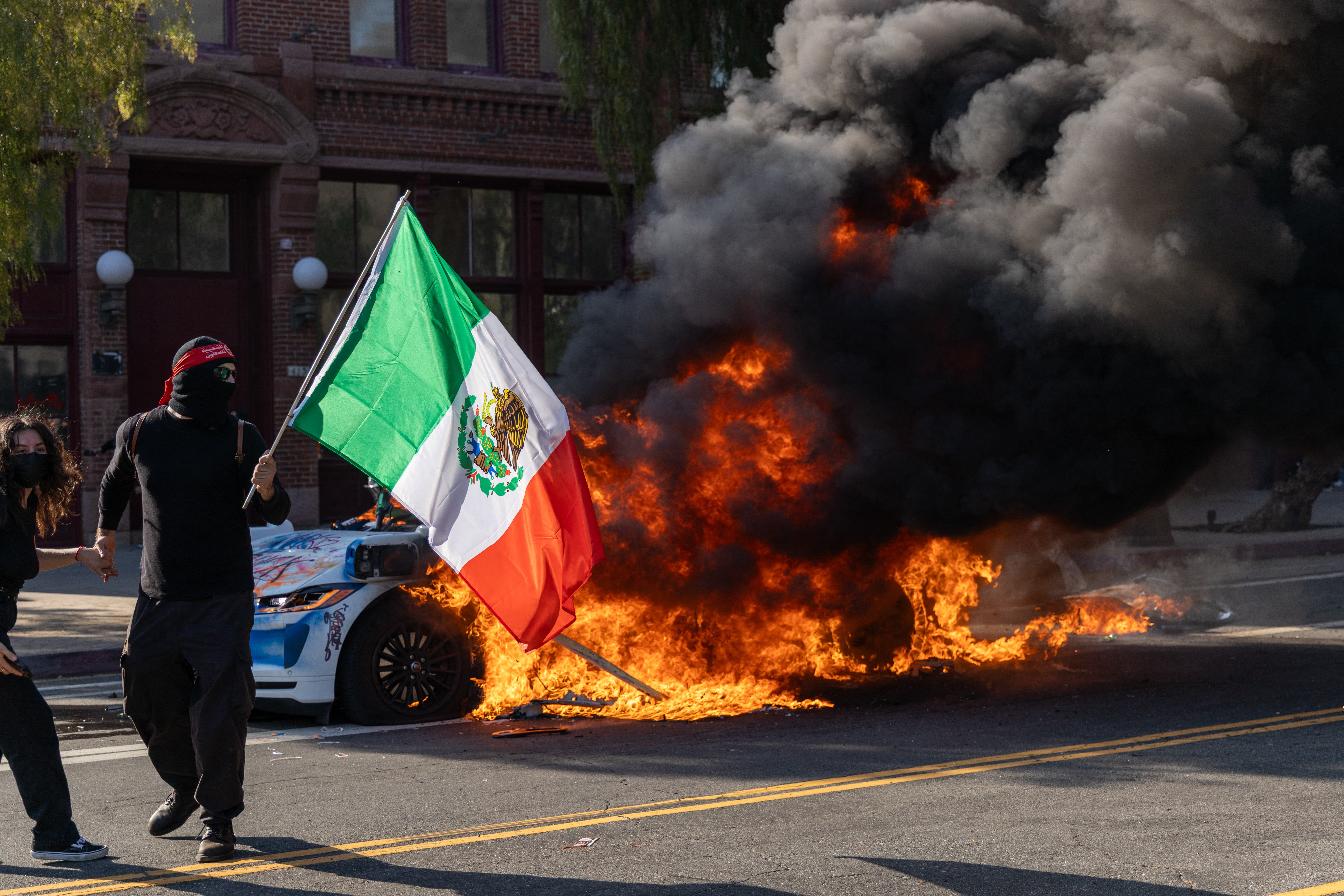 A demonstrator waves a Mexican flag in front of a burning vehicle during a protest in Los Angeles