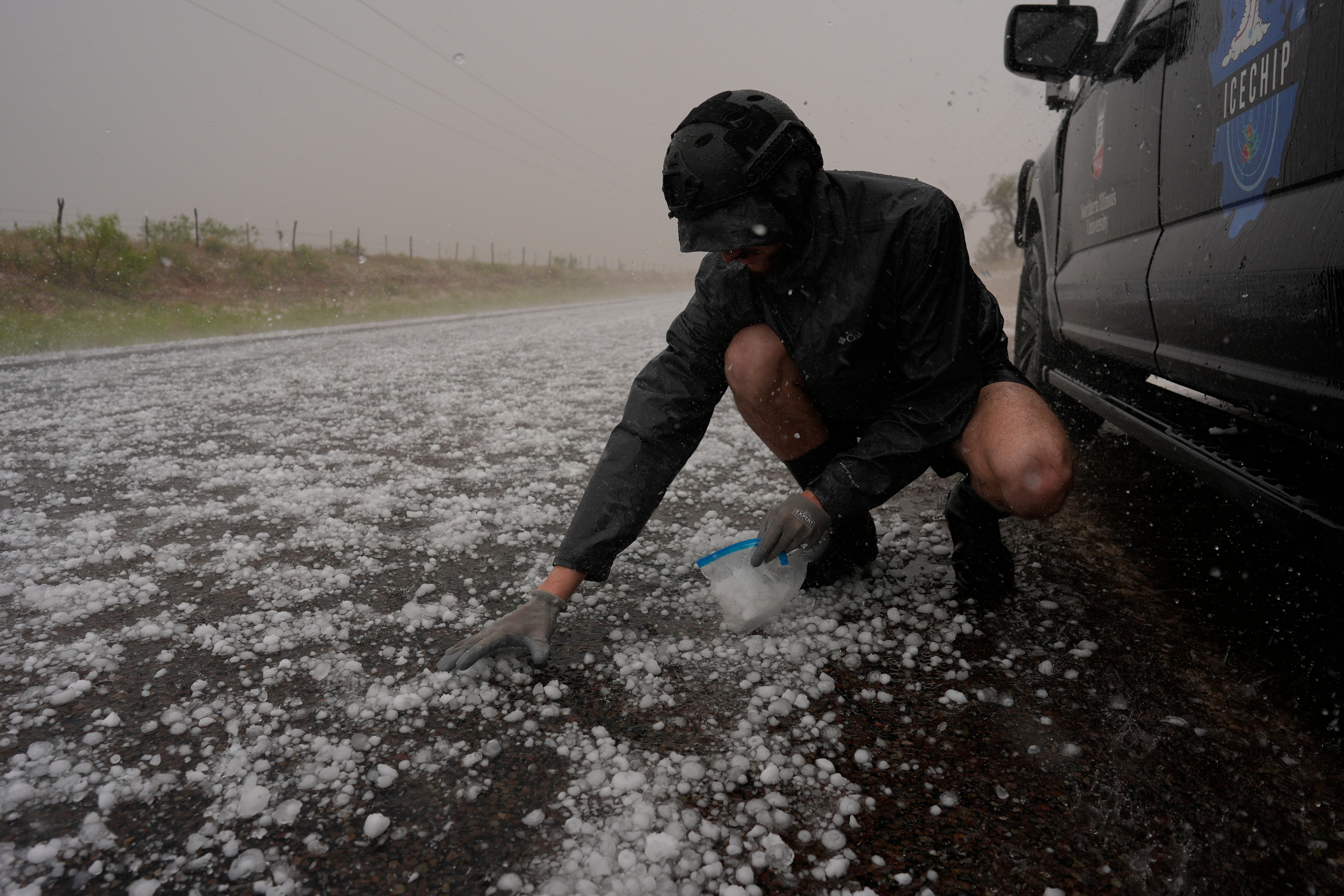 Tony Illenden crouches in a helmet and gloves outside Northern Illinois University's Husky Hail Hunter vehicle to scoop hail into a bag during a storm while on a Project ICECHIP operation Friday, June 6, 2025, in Levelland, Texas