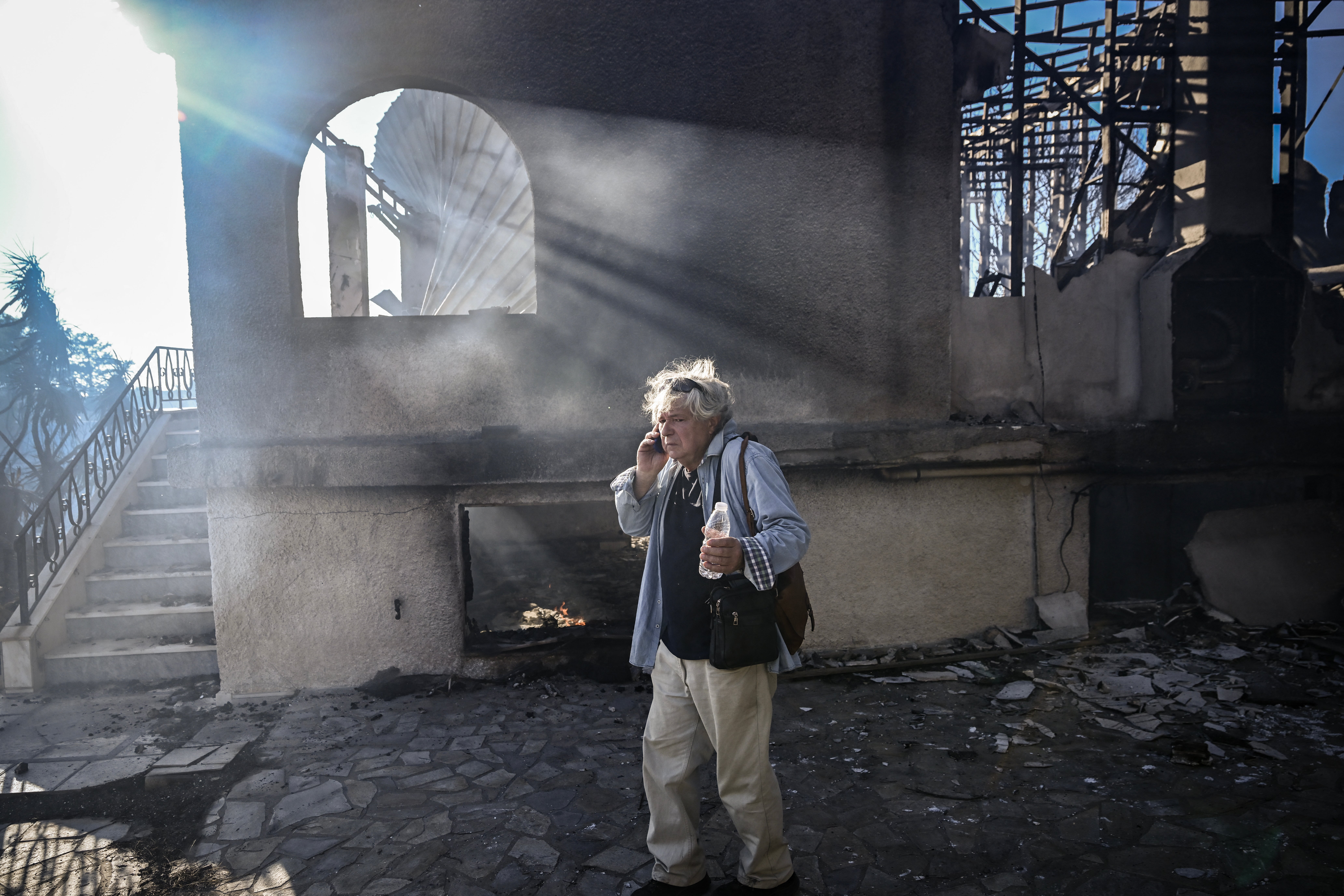 A man talks on his phone as he stands next to a burnt house following a wildfire, in Thymari, southeast of Athens, on June 26, 2025. A forest fire broke out on June 26, 2025 near the seaside towns of Palaia Fokaia and Thymari, 50 km southeast of Athens, leading to evacuations and damaging houses, according to the Greek fire department and public broadcaster Ert. (Photo by Aris MESSINIS / AFP) (Photo by ARIS MESSINIS/AFP via Getty Images)