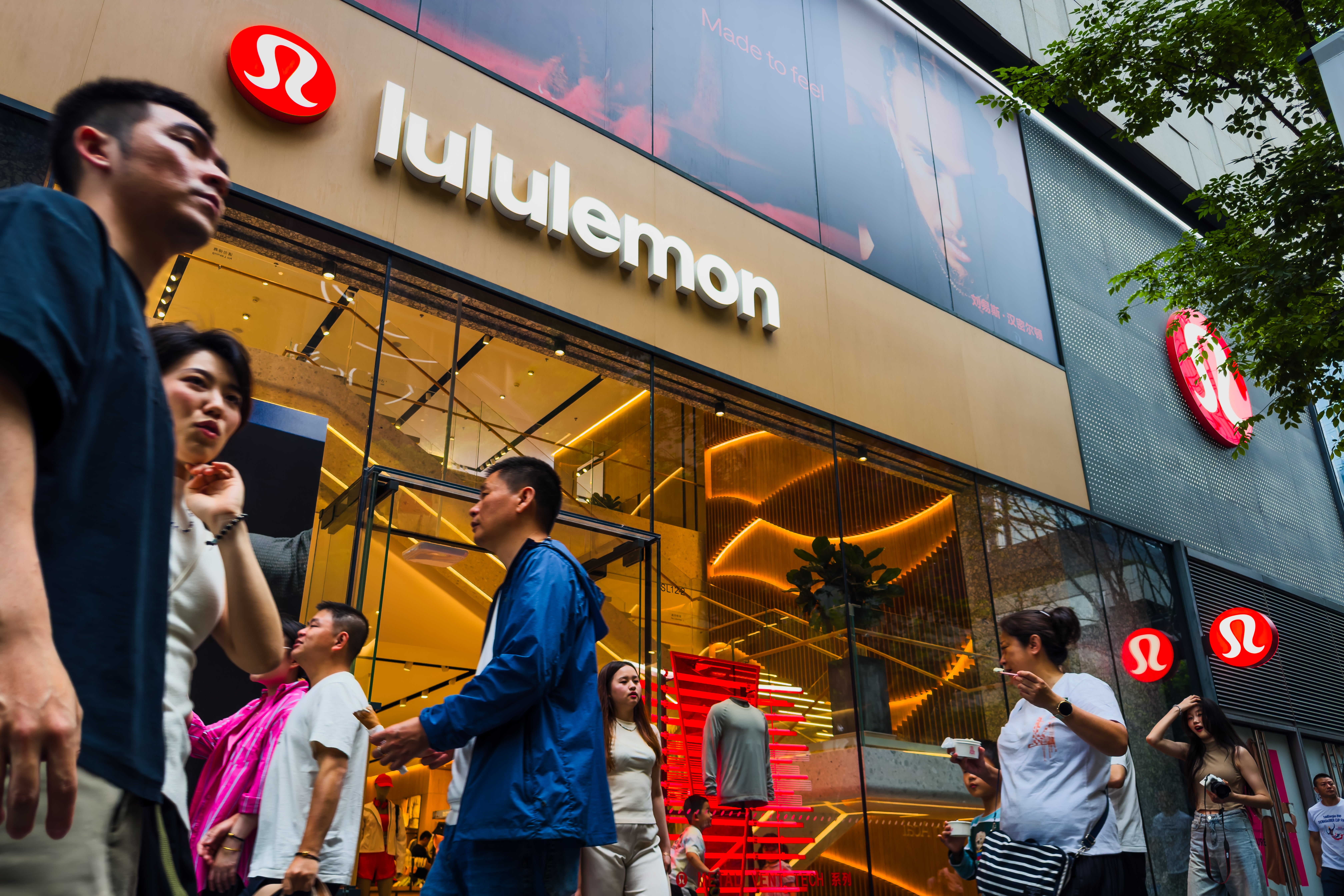 Pedestrians walk past a Lululemon retail store with its brand signage and interior displays visible through large glass windows, while several shoppers are seen entering and exiting the store