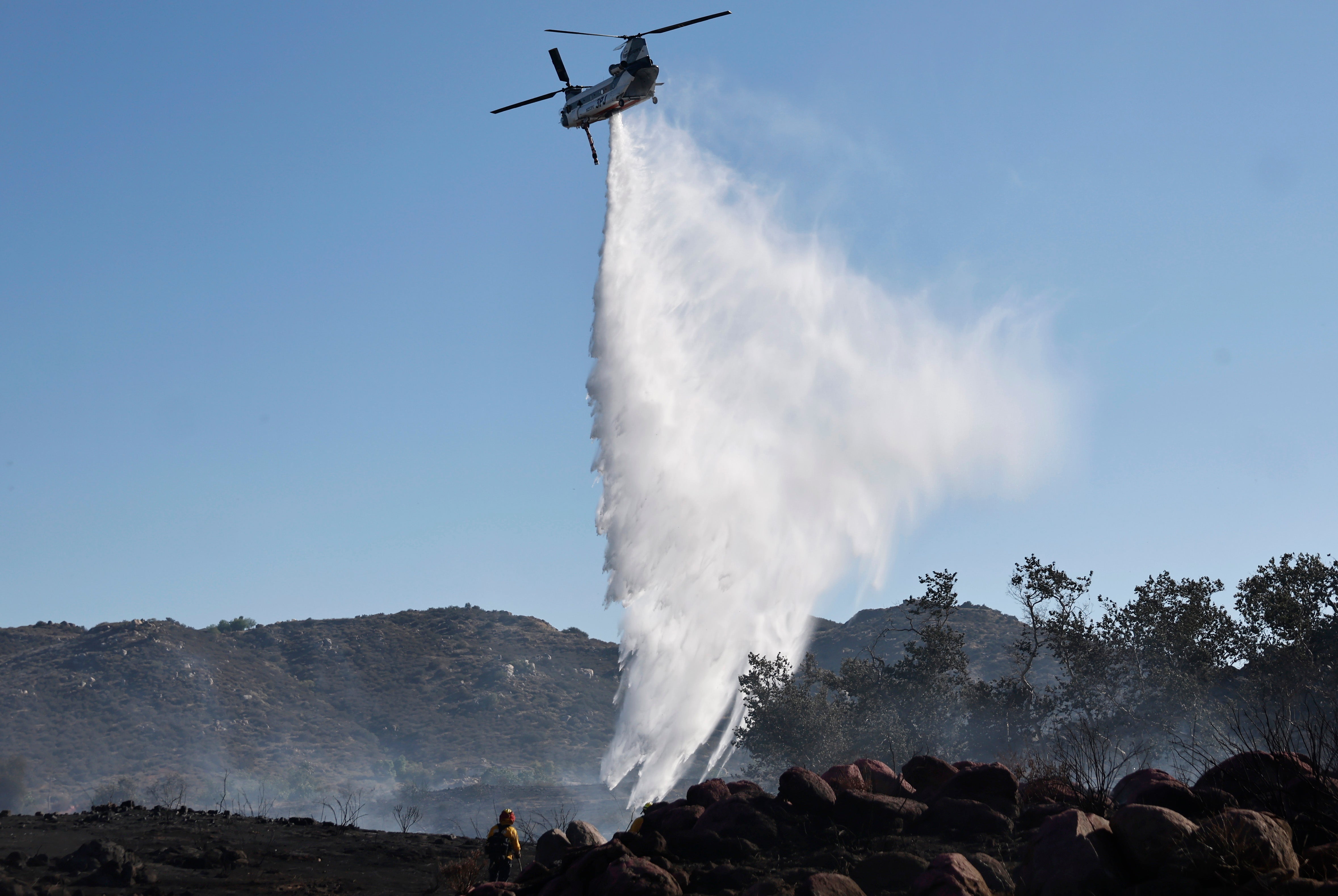 A firefighting helicopter drops water as a firefighter (bottom L) works while the Juniper Fire burns on June 30, 2025