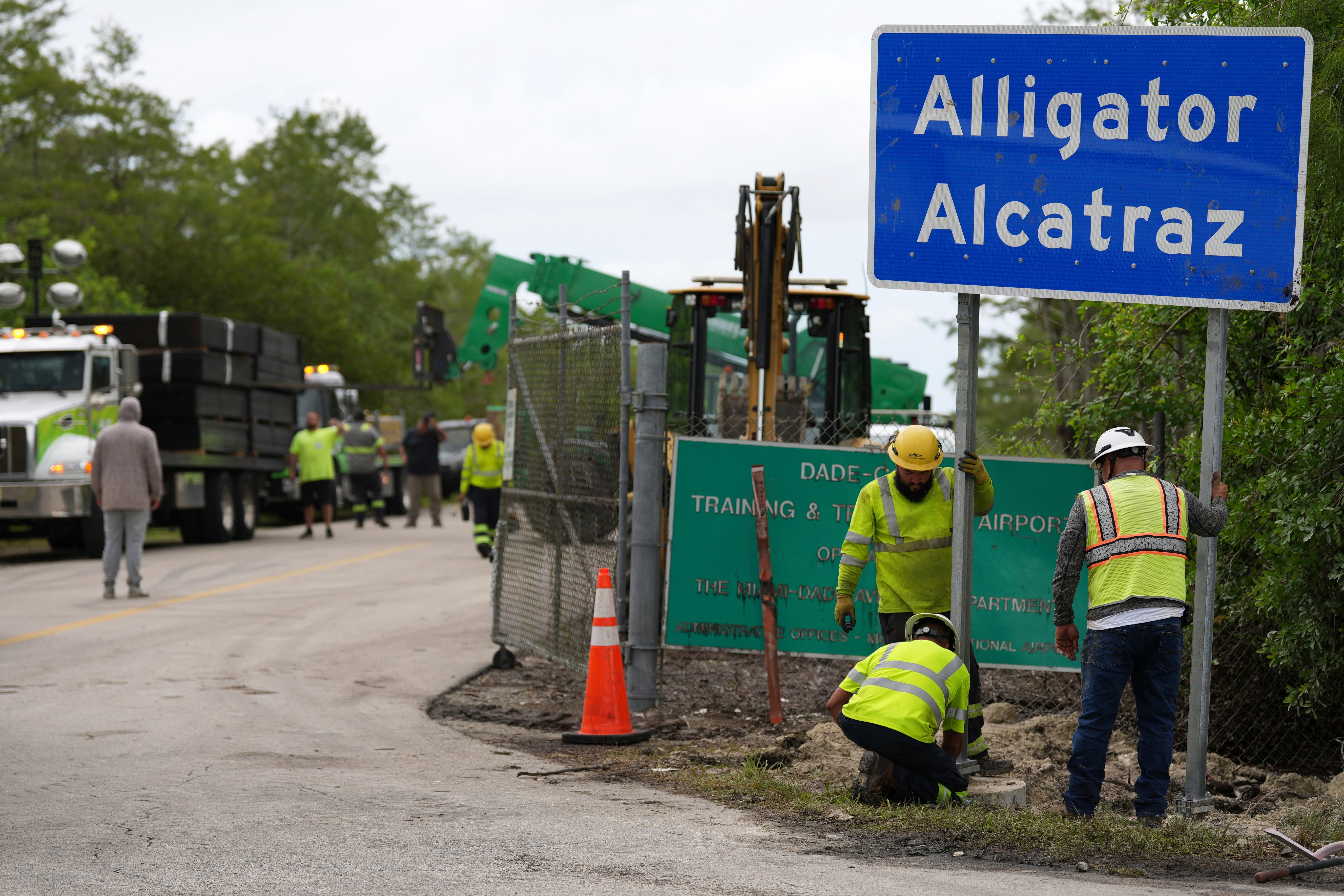 Workers install a sign reading "Alligator Alcatraz" at the entrance to a new migrant detention facility at Dade-Collier Training and Transition facility, as large fencing panels are unloaded from a nearby flatbed, Thursday, July 3, 2025, in Ochopee, Fla.