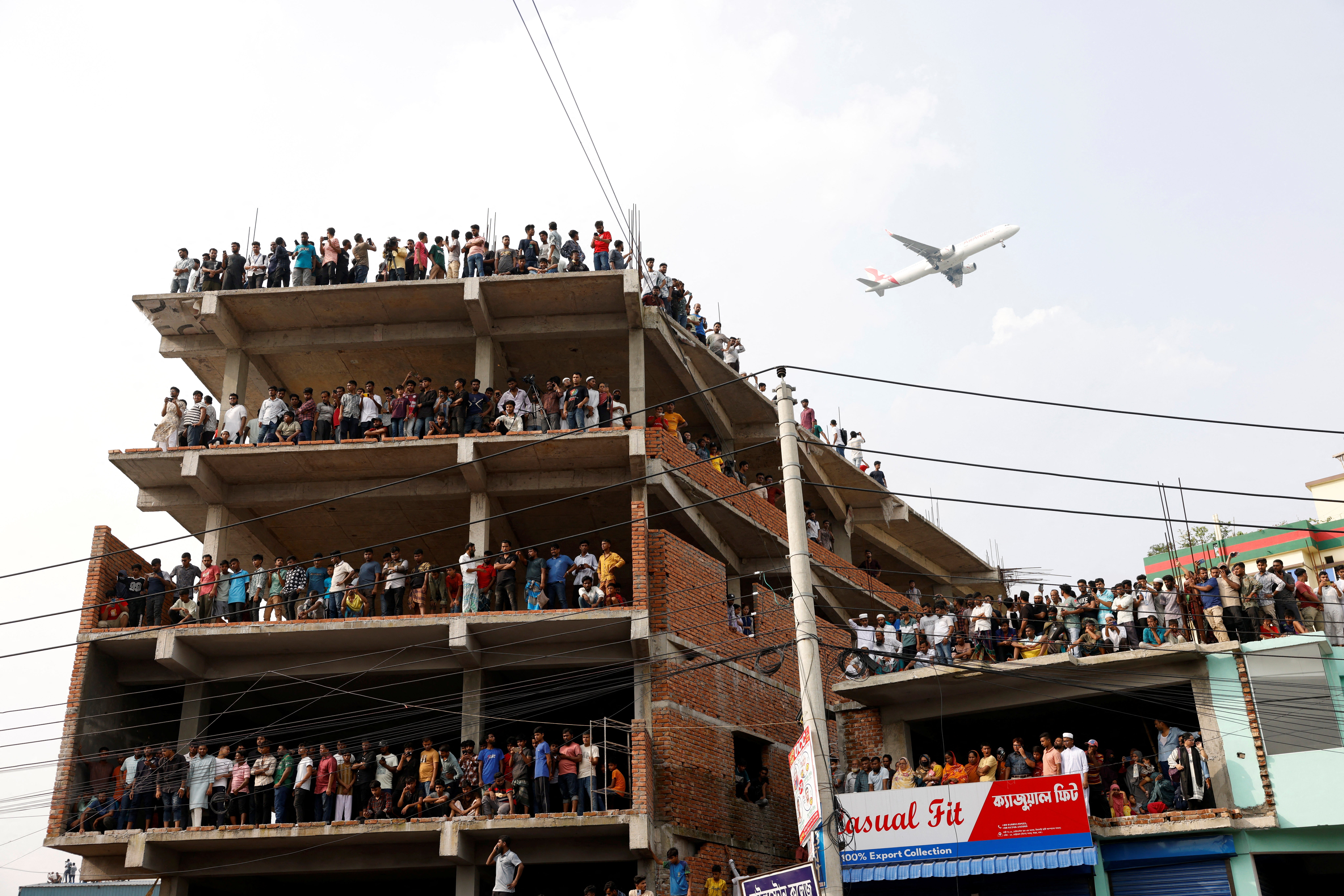 A passenger airplane flies overhead as people watch a rescue operation after an air force aircraft crashed in Dhaka on 21 July 2025