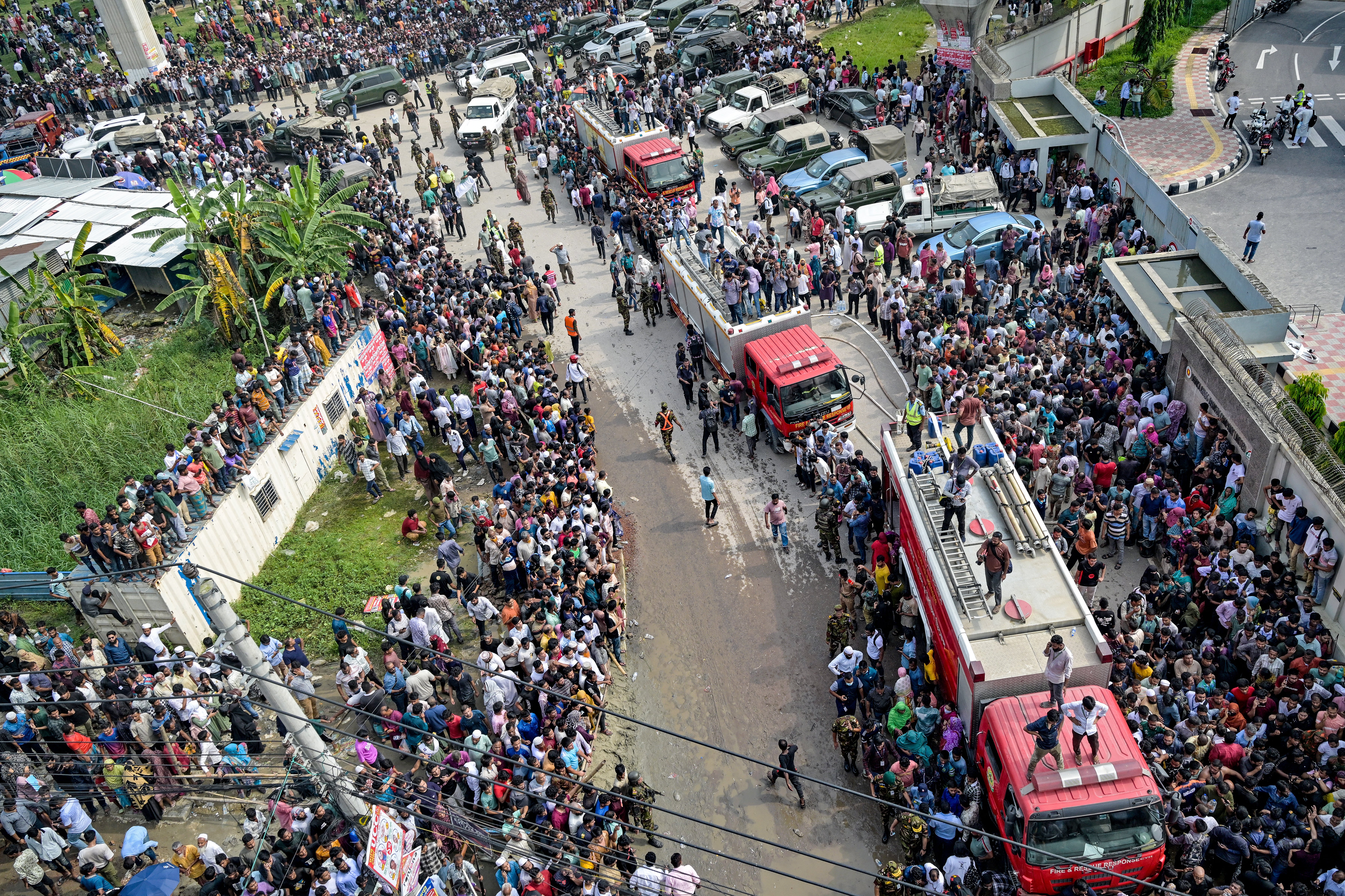 People crowd a street as fire fighting trucks remain on standby outside a school where an Air Force training jet crashed in Dhaka on Monday 21 July 2025