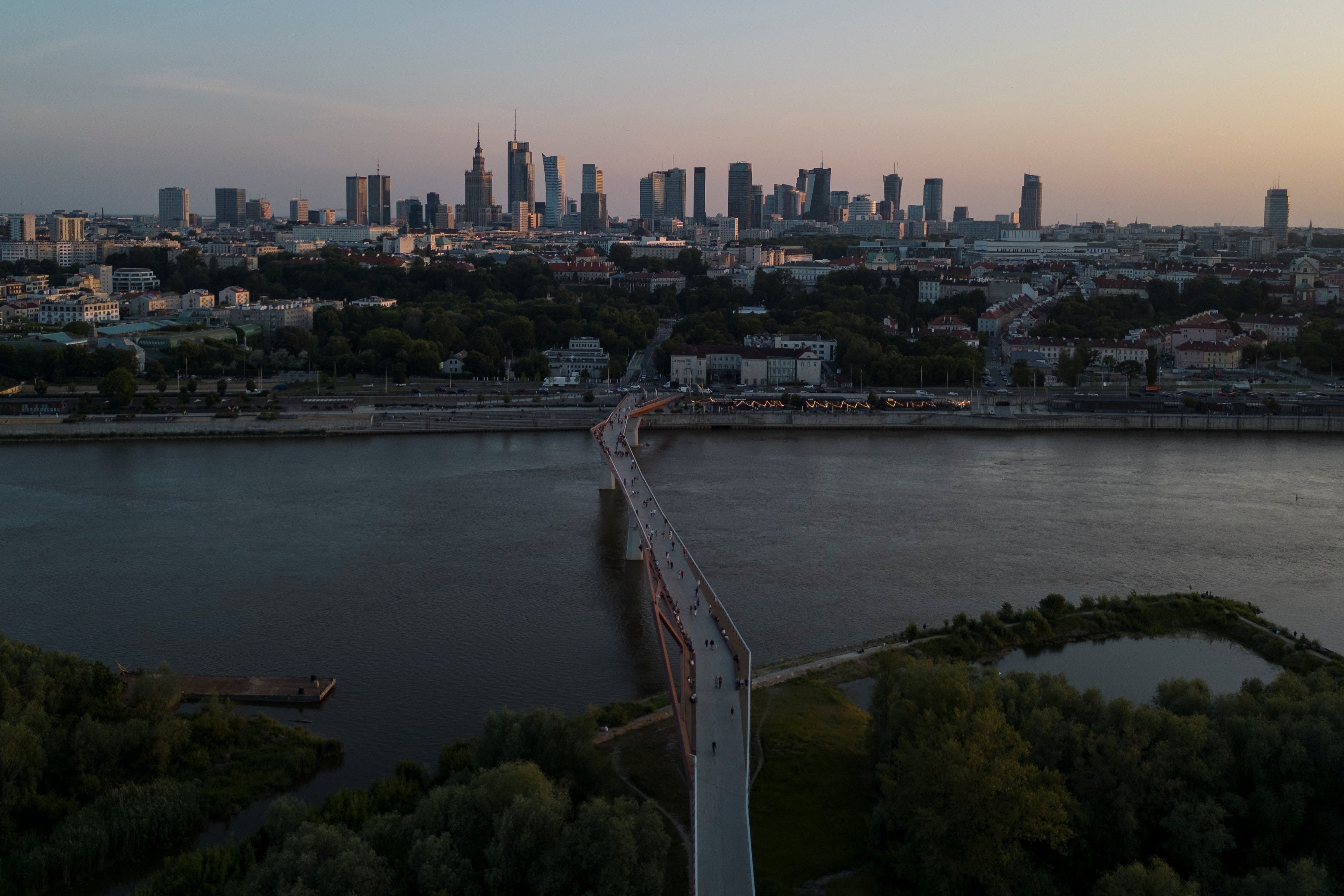 A drone view shows the skyline of Warsaw with the Vistula River and a pedestrian footbridge in the foreground, and the Palace of Culture and Science visible among the city’s landmarks in the background