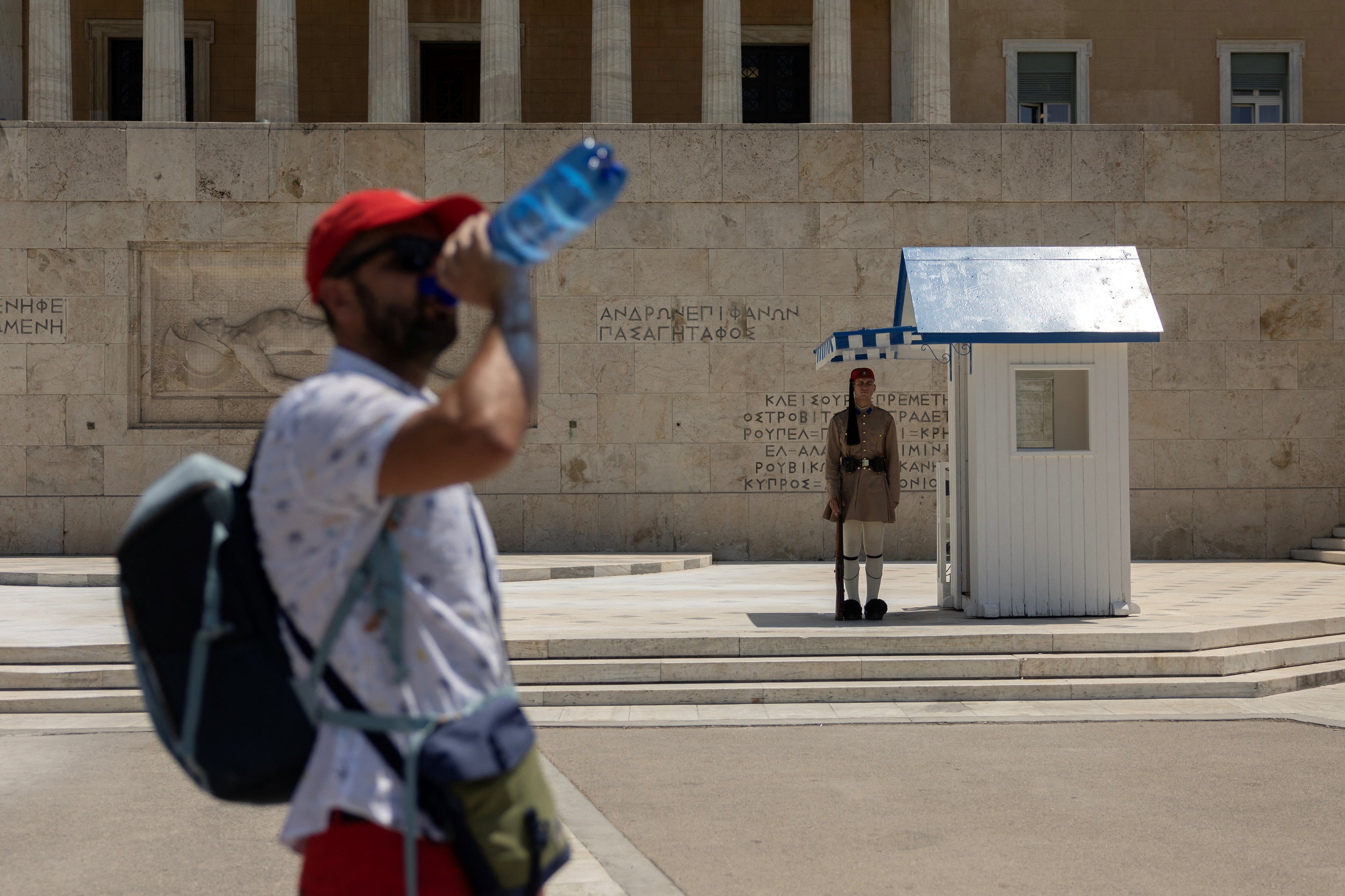 A person drinks water with a member of Greek Presidential Guard in the background, in front of the parliament building, during a heatwave in Athens