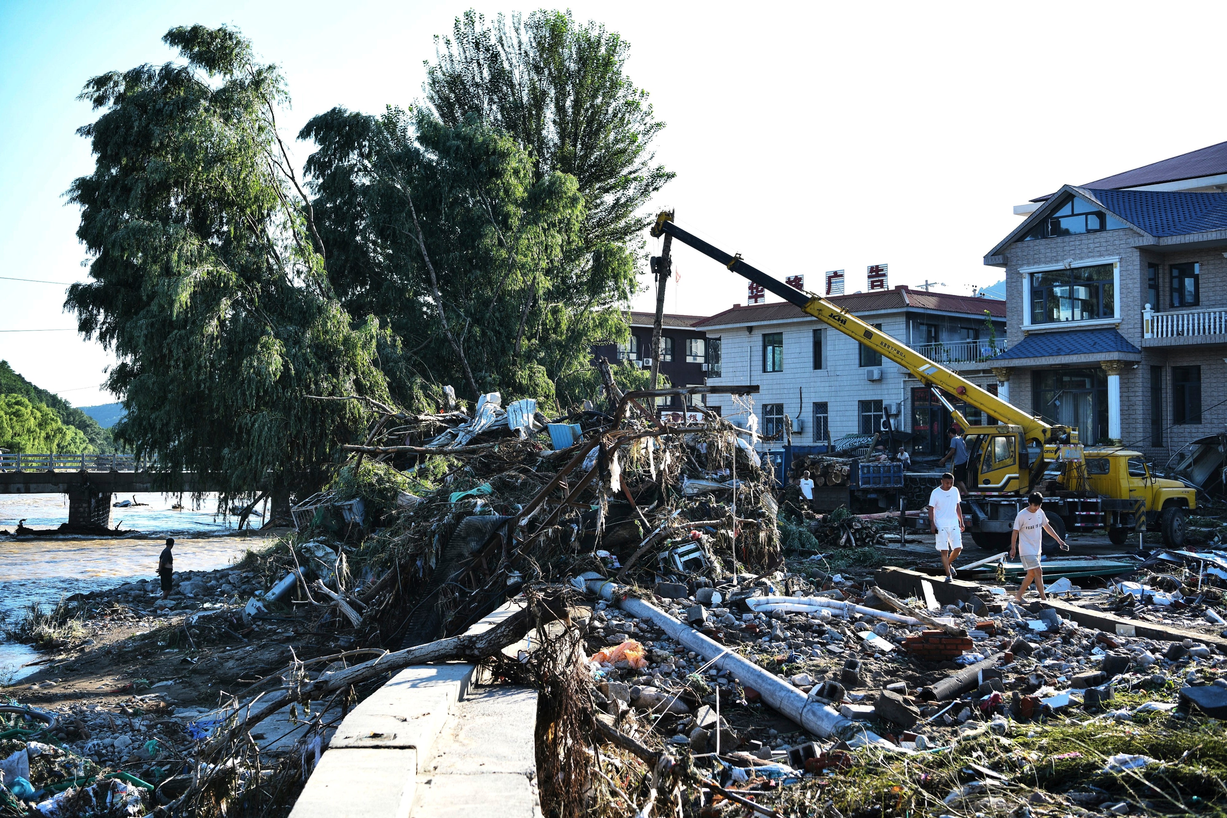 People walk past workers clearing debris along a damaged riverbank after a flash flood in Jizhou district of northern China's Tianjin municipality
