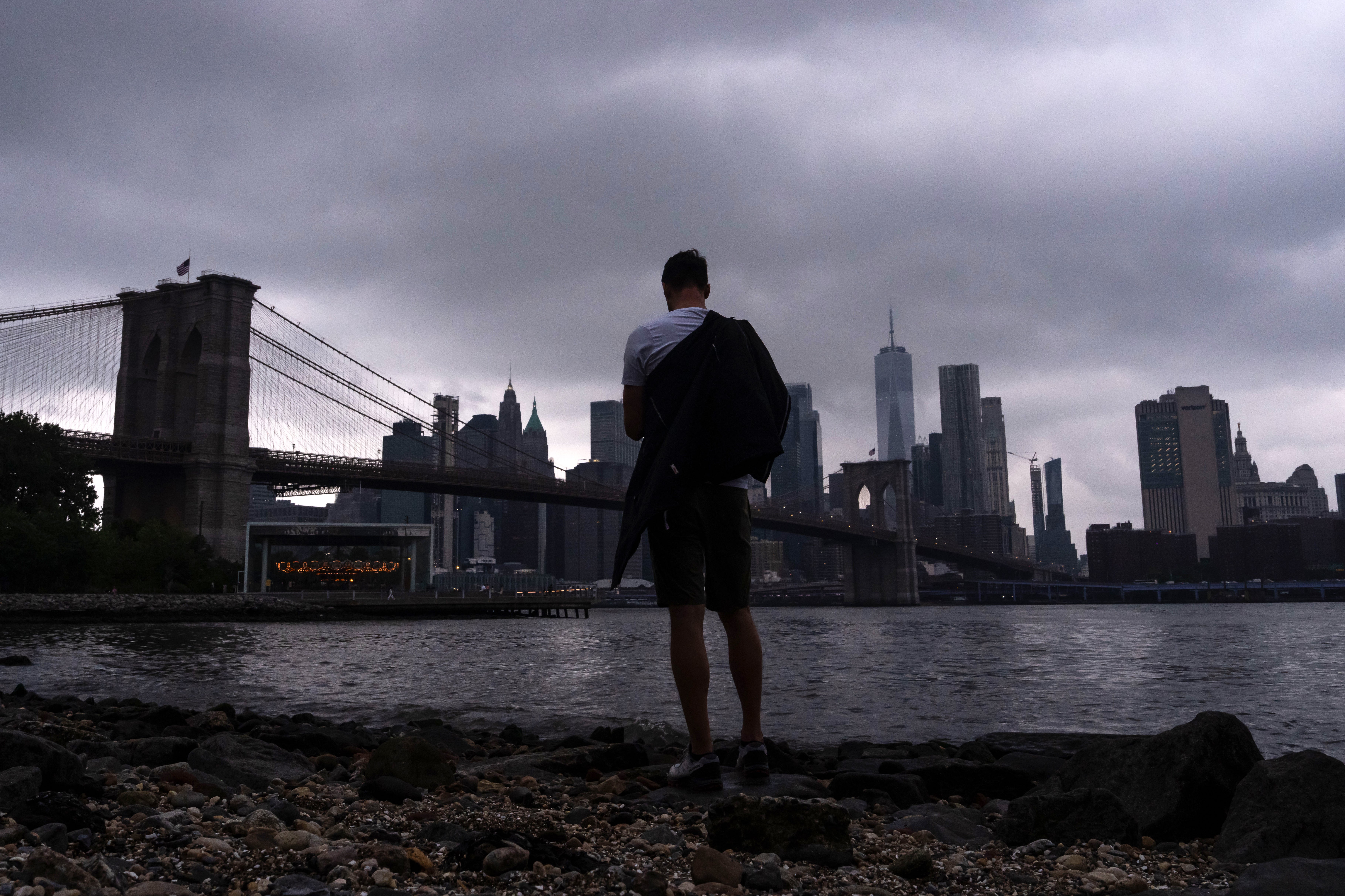 A person stands by the East River in front of the Brooklyn Bridge during a break in the rain on July 31, 2025 in the Brooklyn borough of New York City