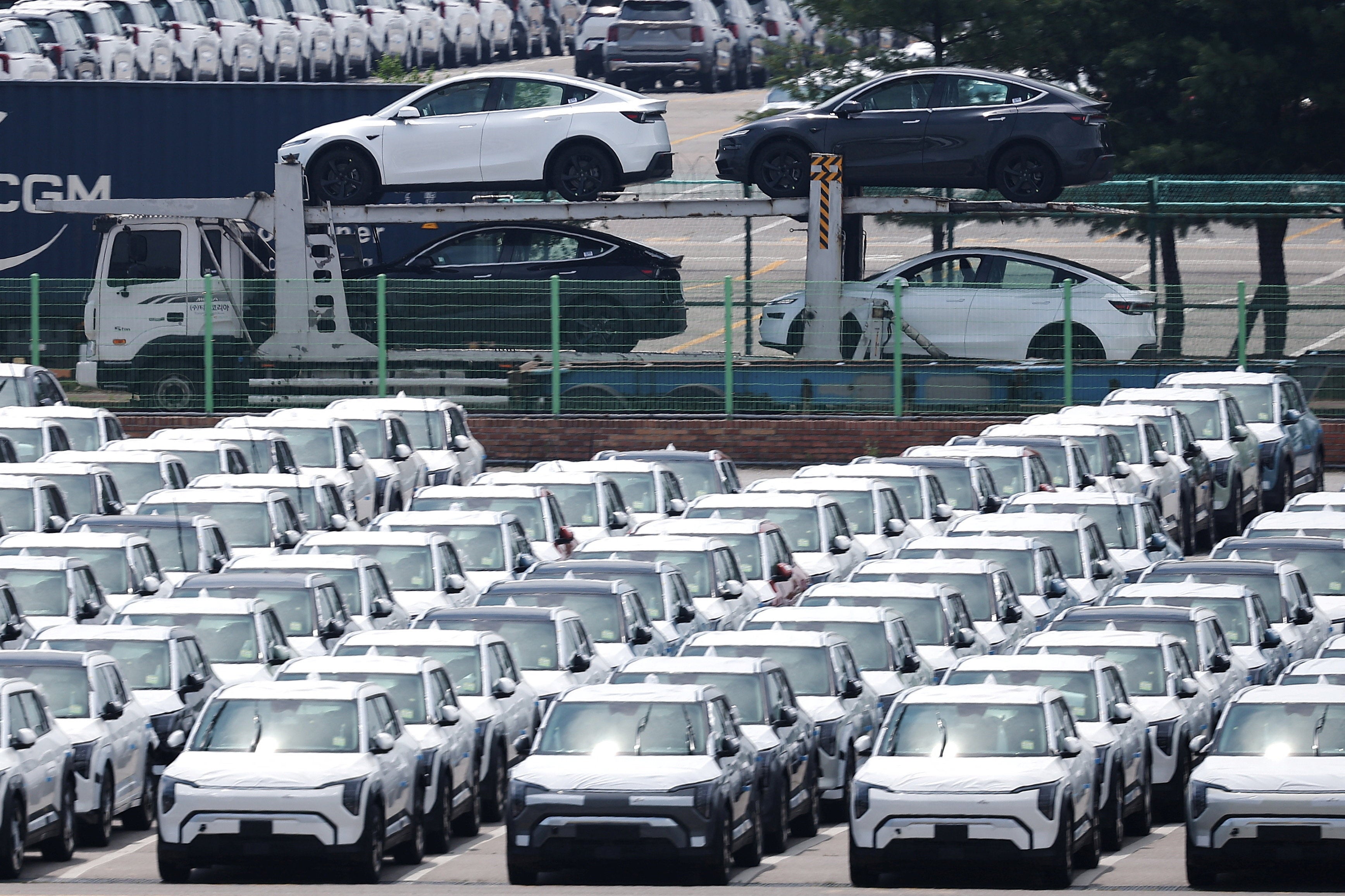 A carrier transports Tesla vehicles travels past KIA vehicles parked at a port in Pyeongtaek
