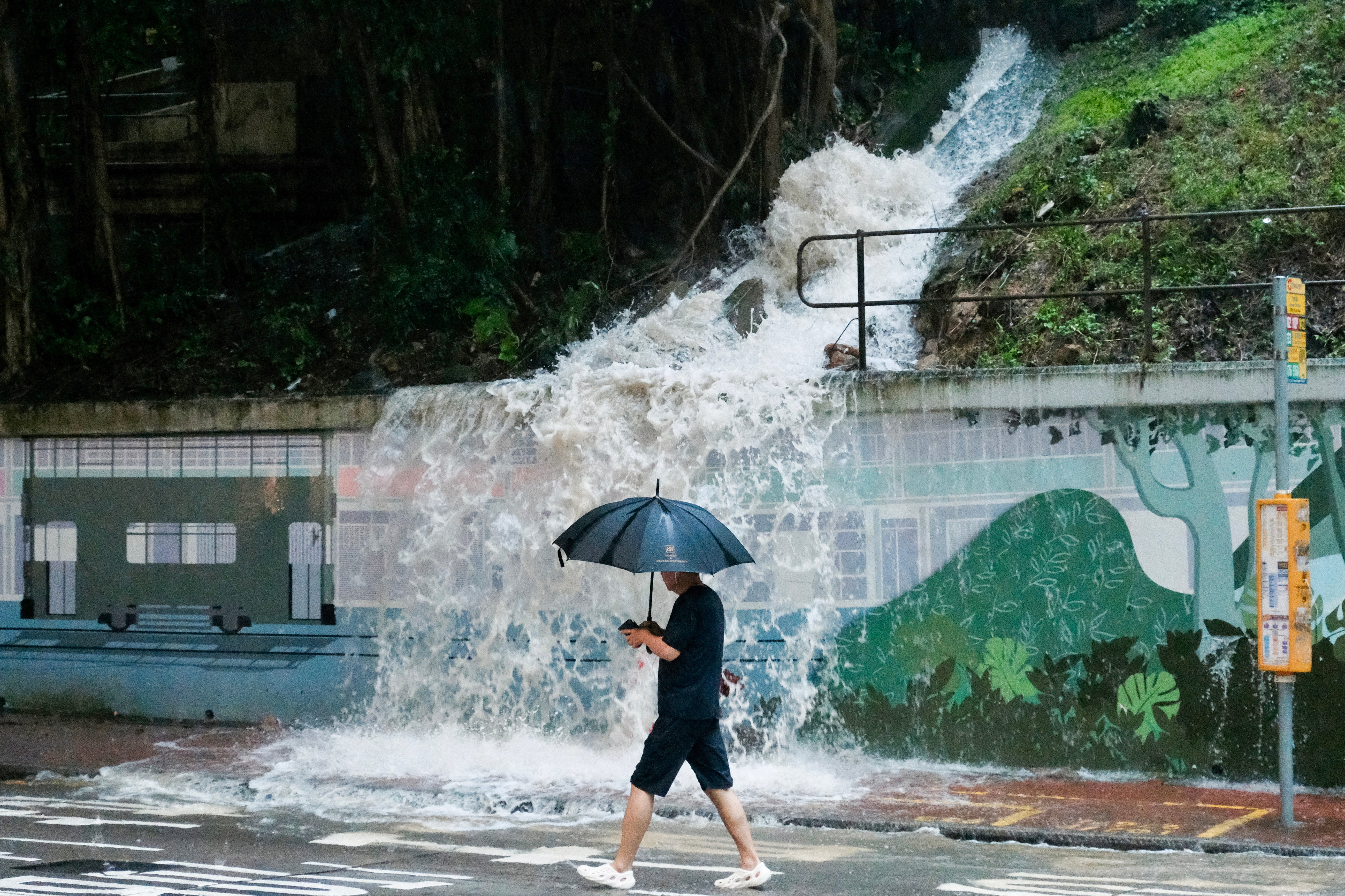 Pedestrian walks past an overflowing hillside drain spilling water onto a street in the Quarry Bay area of Hong Kong on 5 August 2025