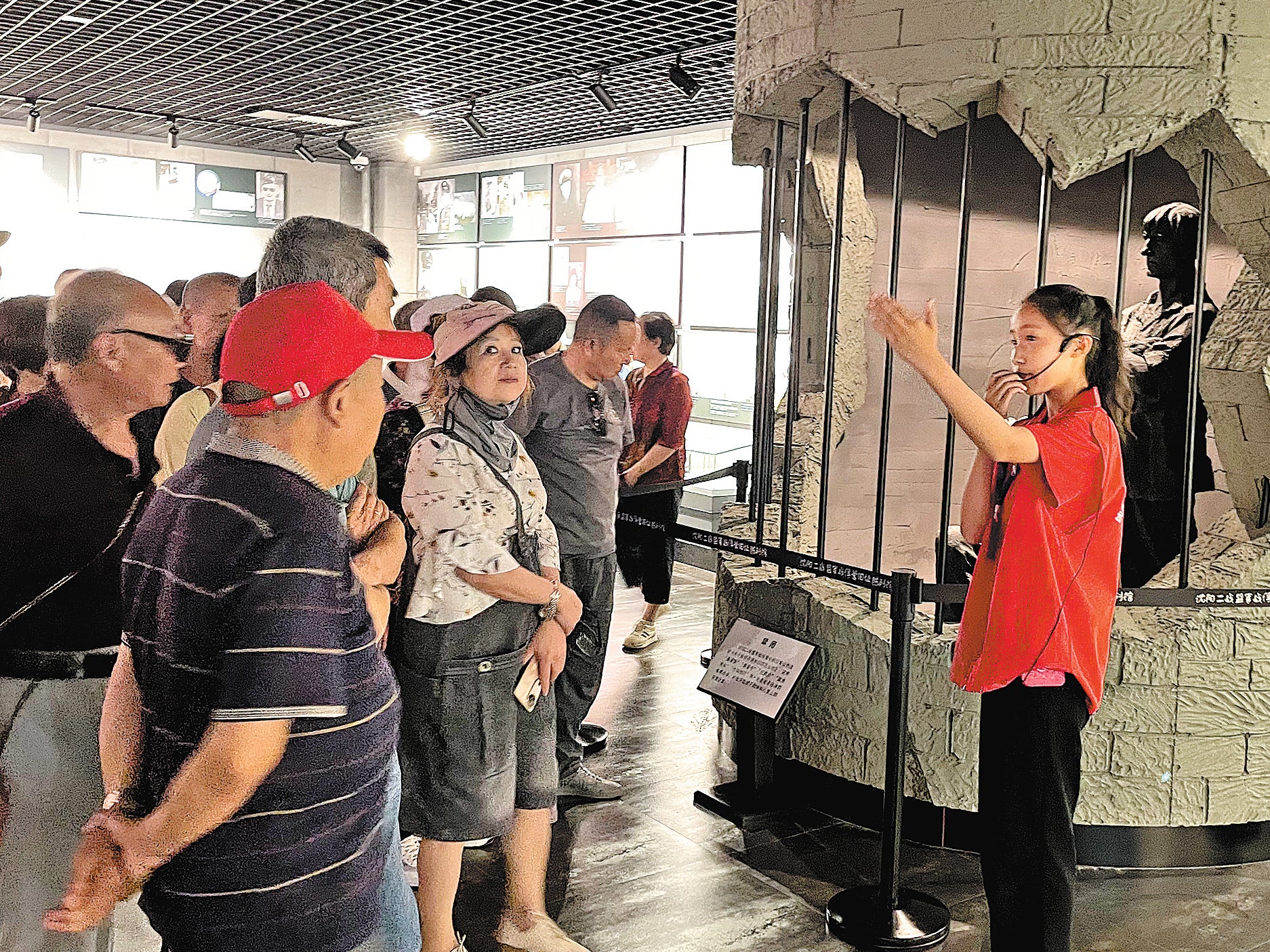 At the Shenyang WWII Allied Prisoners Camp Site Museum, a young volunteer guide addresses visitors. In the background is a model depicting a brutal punishment method, in which POWs were confined in tiny spaces without food or water