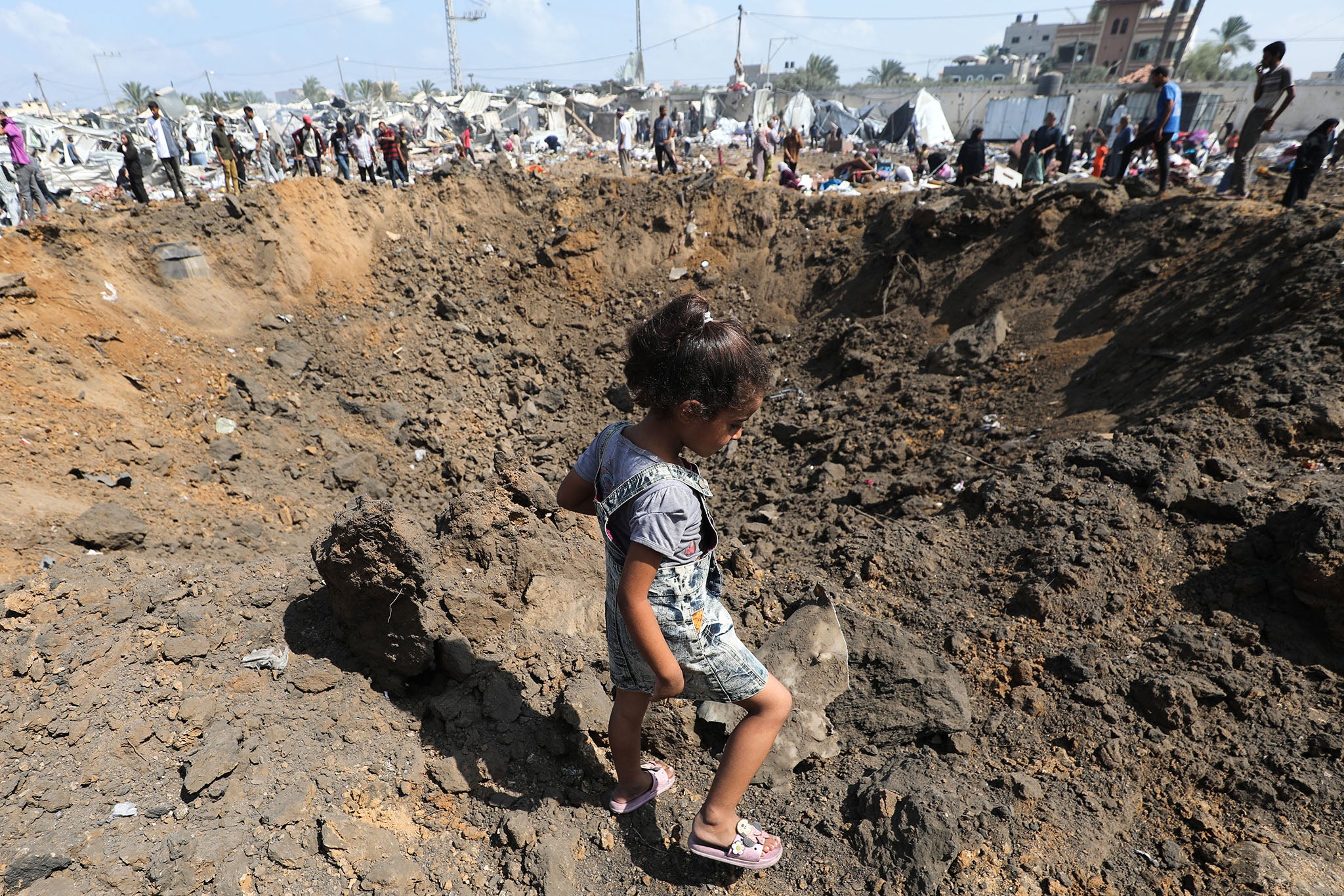 A displaced Palestinian girl walks next to an impact crater, left behind after an Israeli strike, at a camp for internally displaced people in Deir el-Balah in the central Gaza Strip