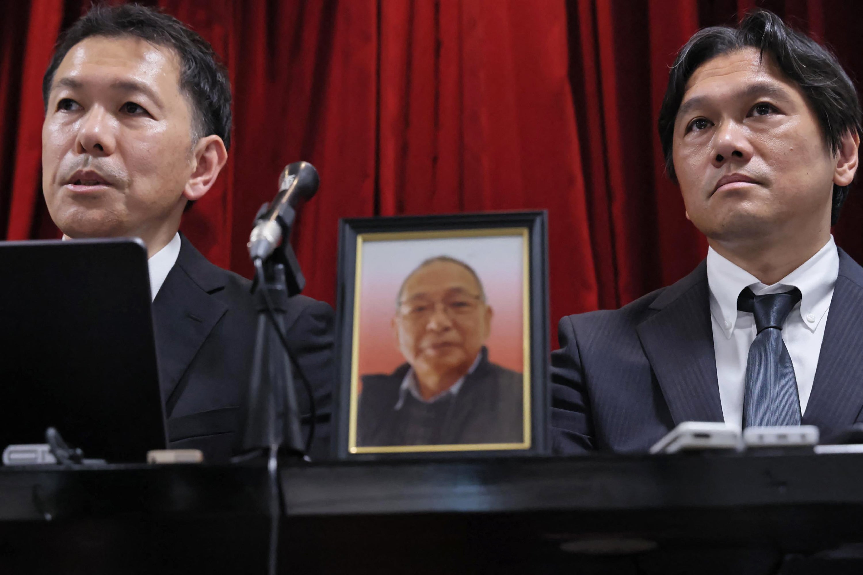 A portrait of the late Shizuo Aishima (C), who was falsely accused of unauthorised export of sensitive equipment and died after prolonged detention, is seen as his sons hold a press conference in Yokohama on 25 August 2025, after police and prosecutors apologised at Aishima's grave for his illegal detention