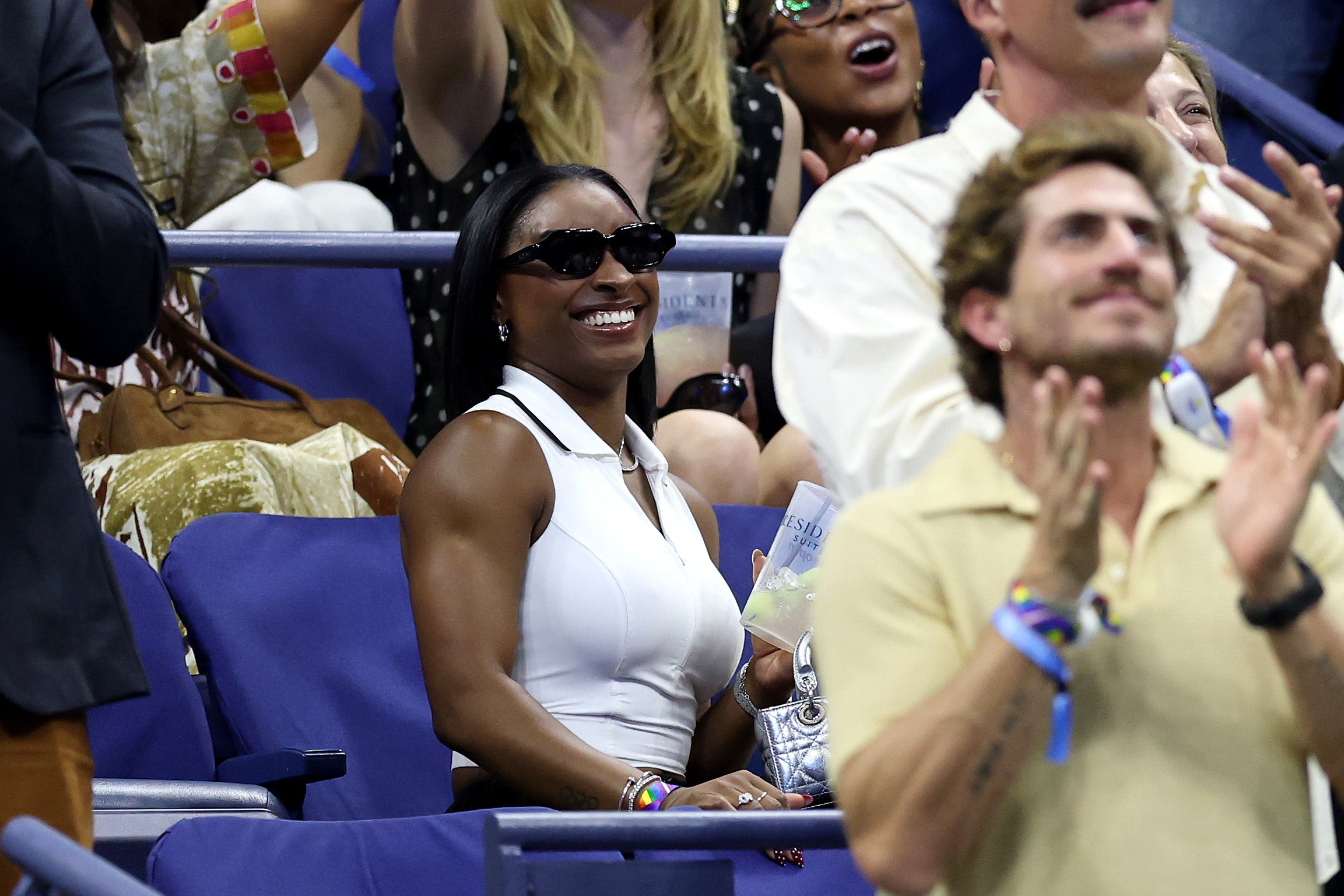 Simone Biles sat in the stands at the US Open in a white, zip-up polo crop top on August 28. She paired the shirt with silver jewelry, sunglasses, and black pants
