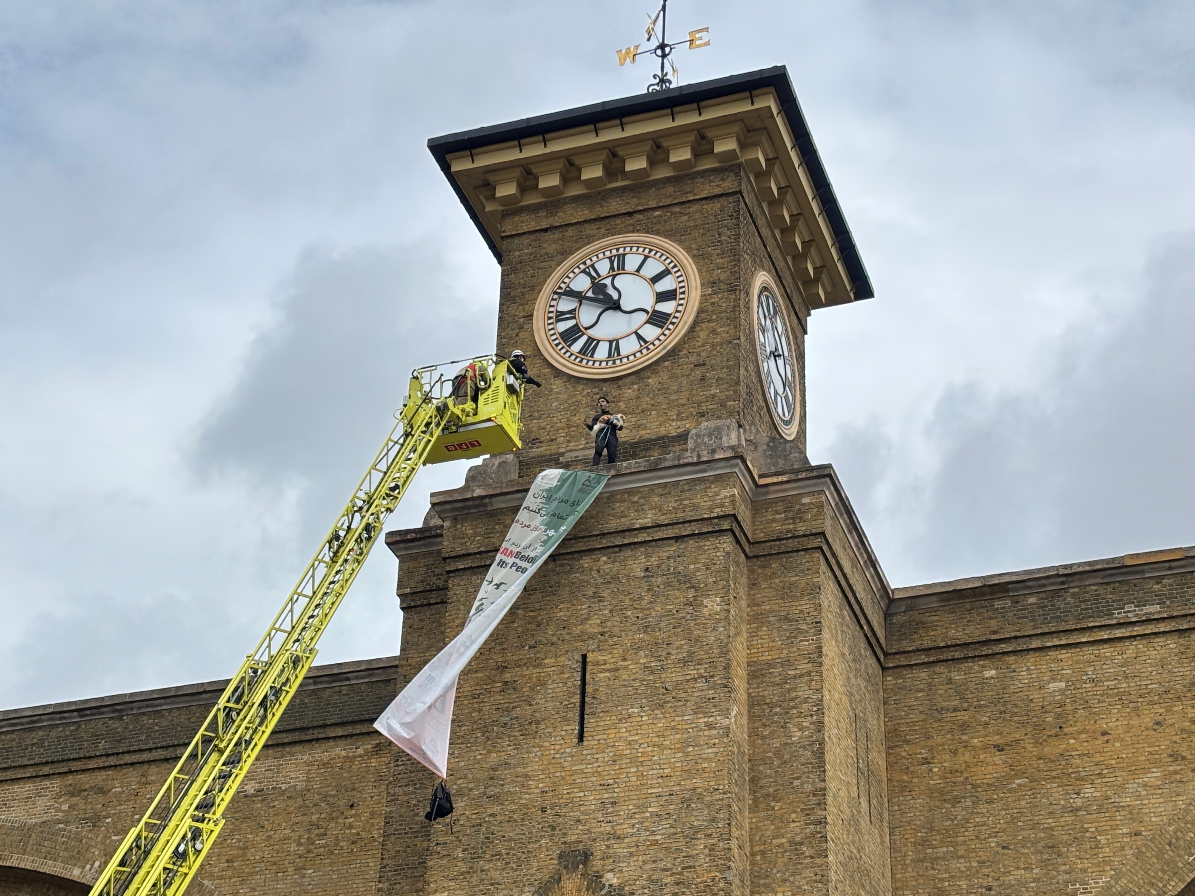 The protester is wearing a black t-shirt which says "no to war", with a banner saying "Iran belongs to its people" draped down the tower at King’s Cross station