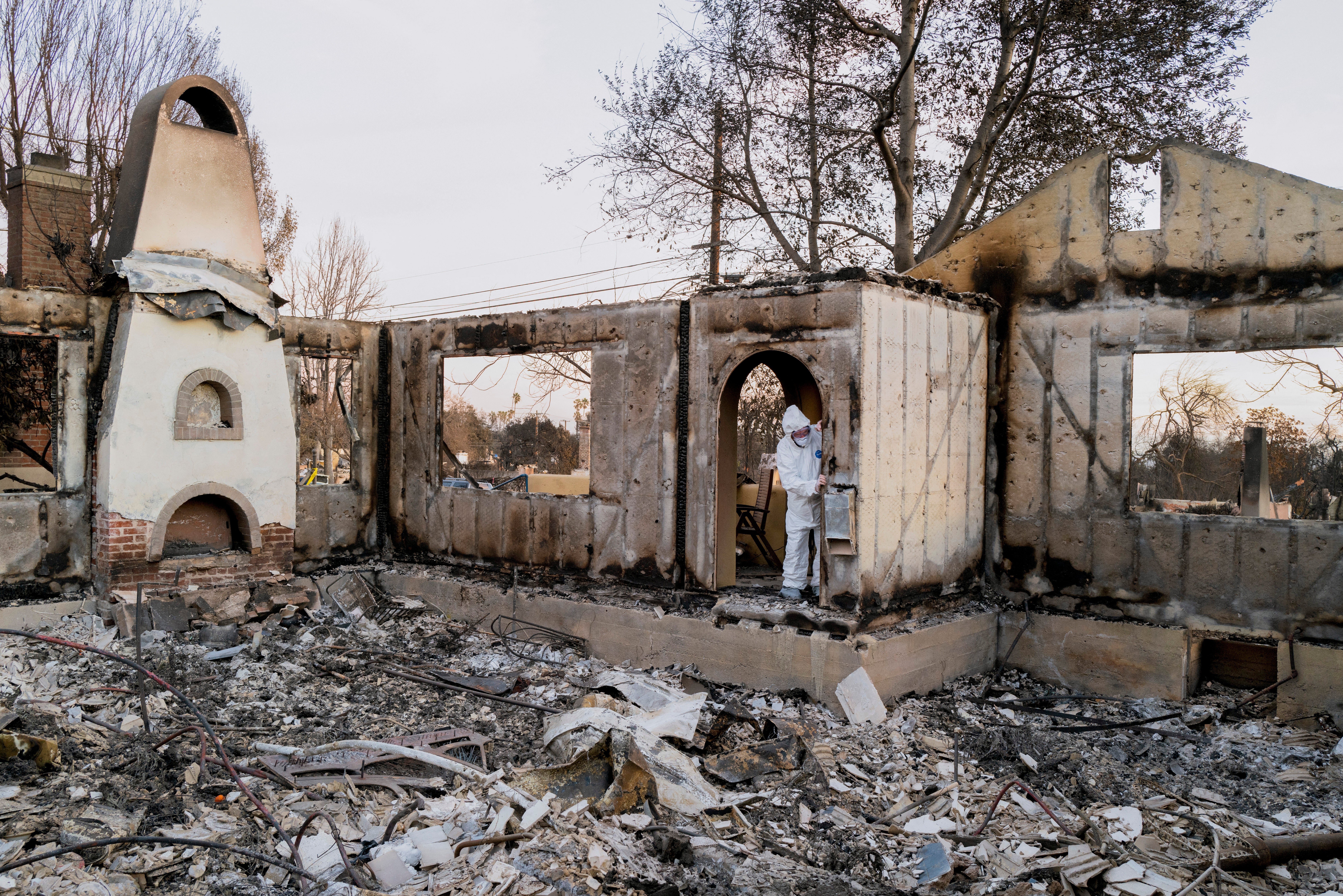 A person in protective gear navigates through the ruins of a burned home in Altadena, the heart of the Eaton Fire