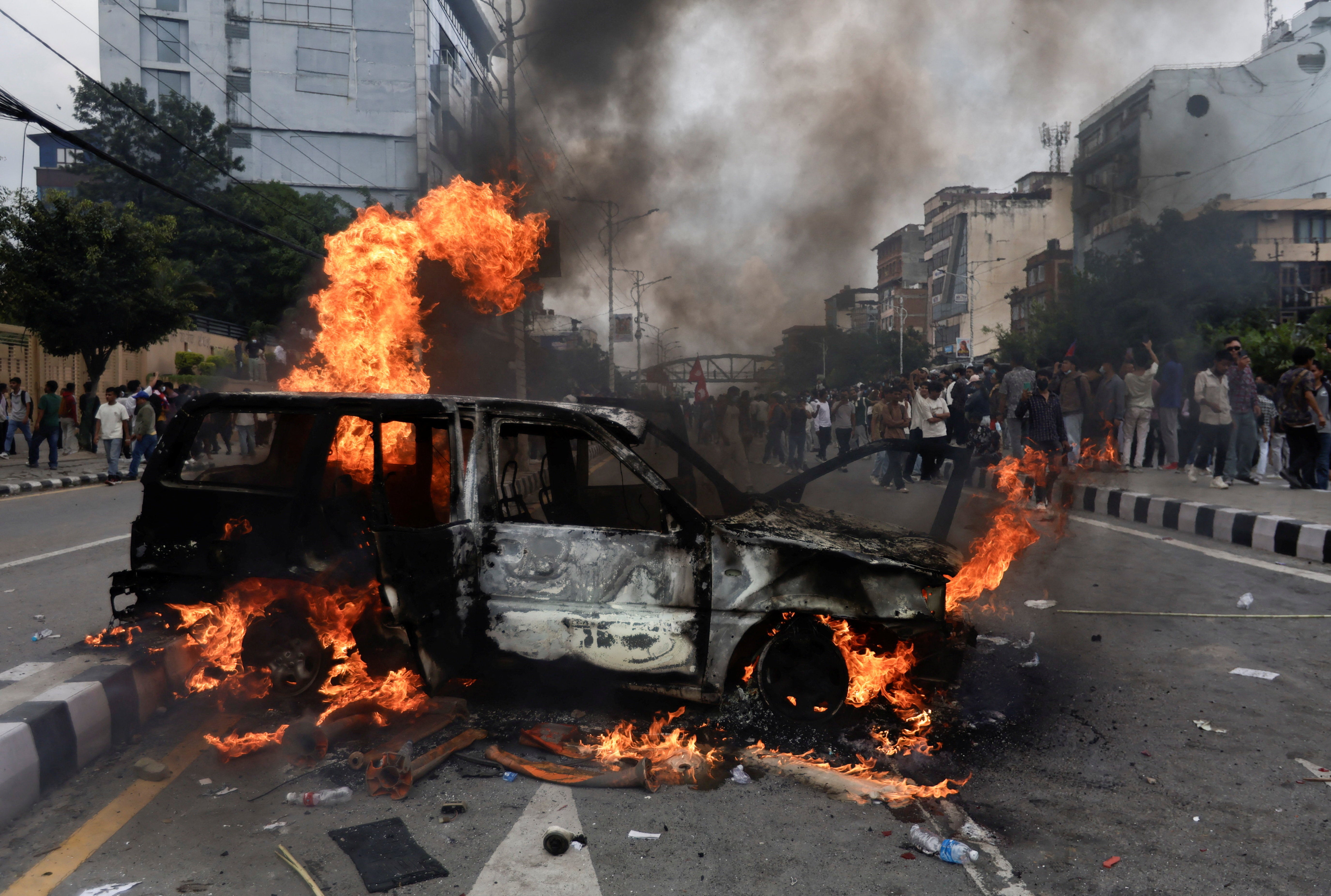 People stand near a vehicle torched by the demonstrators during a protest against corruption and the government's decision to block several social media platforms