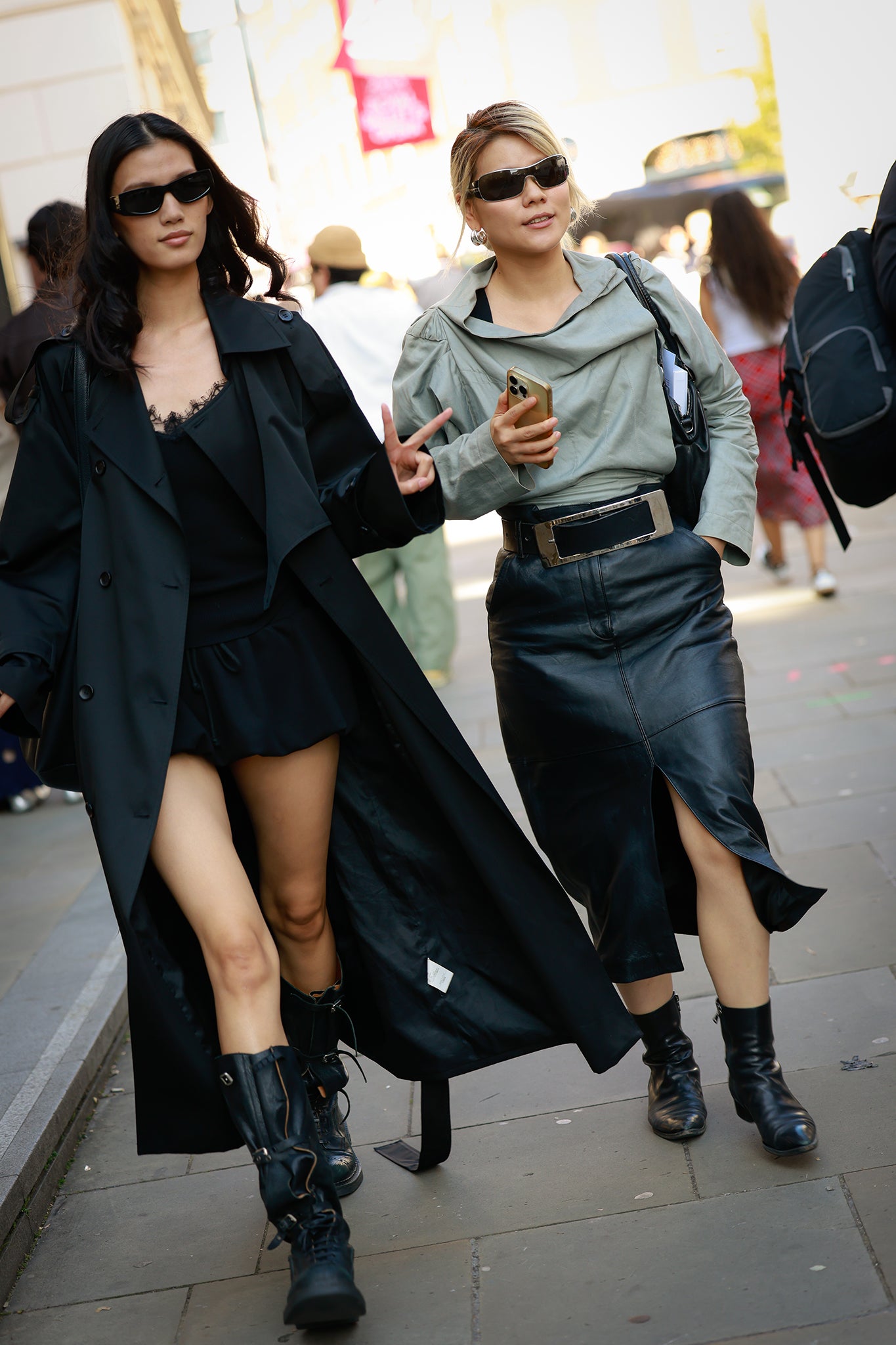 A guest (left) wearing black sunglasses, black trench coat, black lacy top and black boots and a guest wearing grey top, black leather front split midi skirt, black ankle boots and a black shoulder bag outside Mithridate during London Fashion Week on 19 September 2025 (photo by Hanna Lassen/Getty Images)