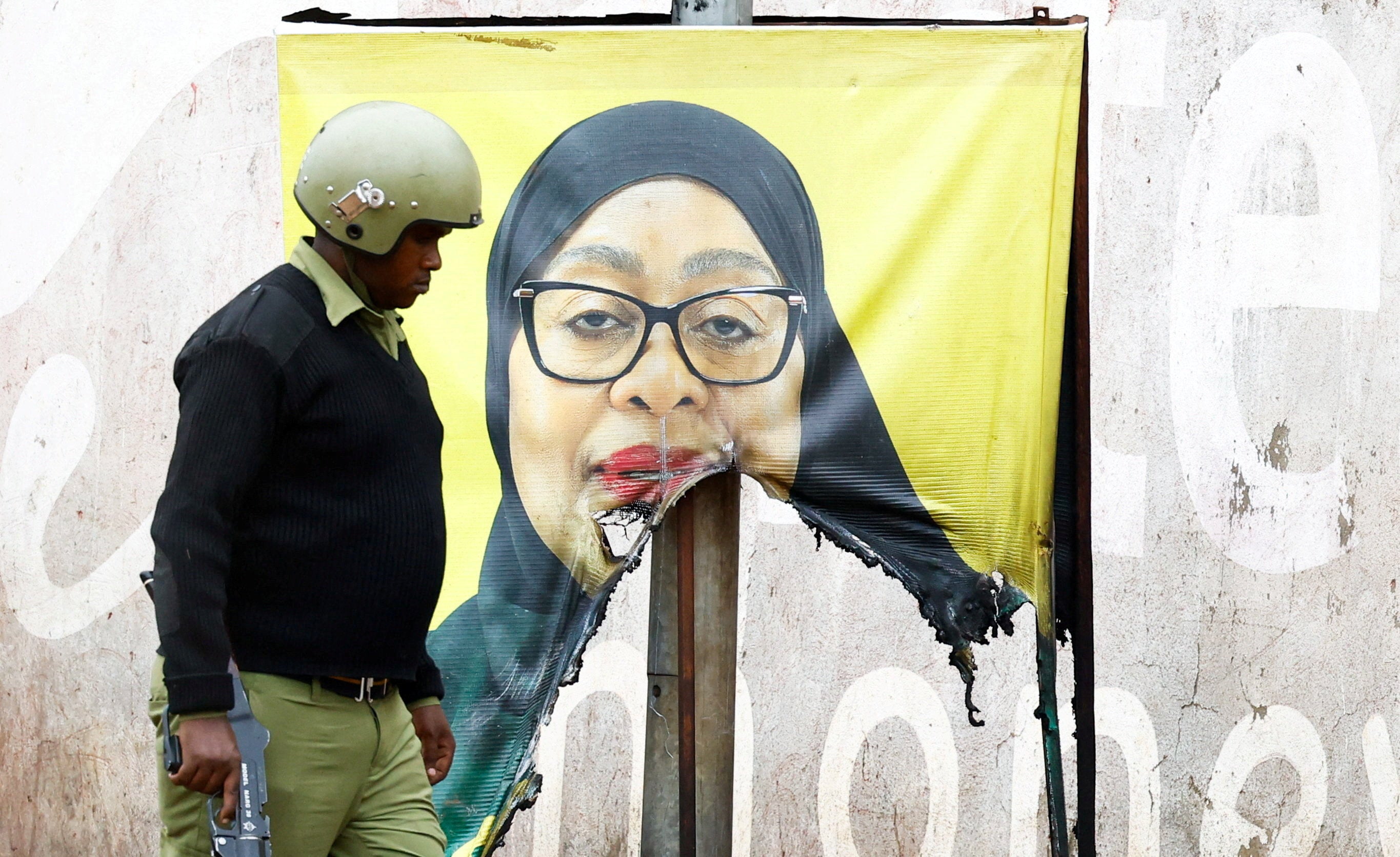 A Tanzanian riot police officer walks past a vandalised campaign poster of President Samia Suluhu Hassan.