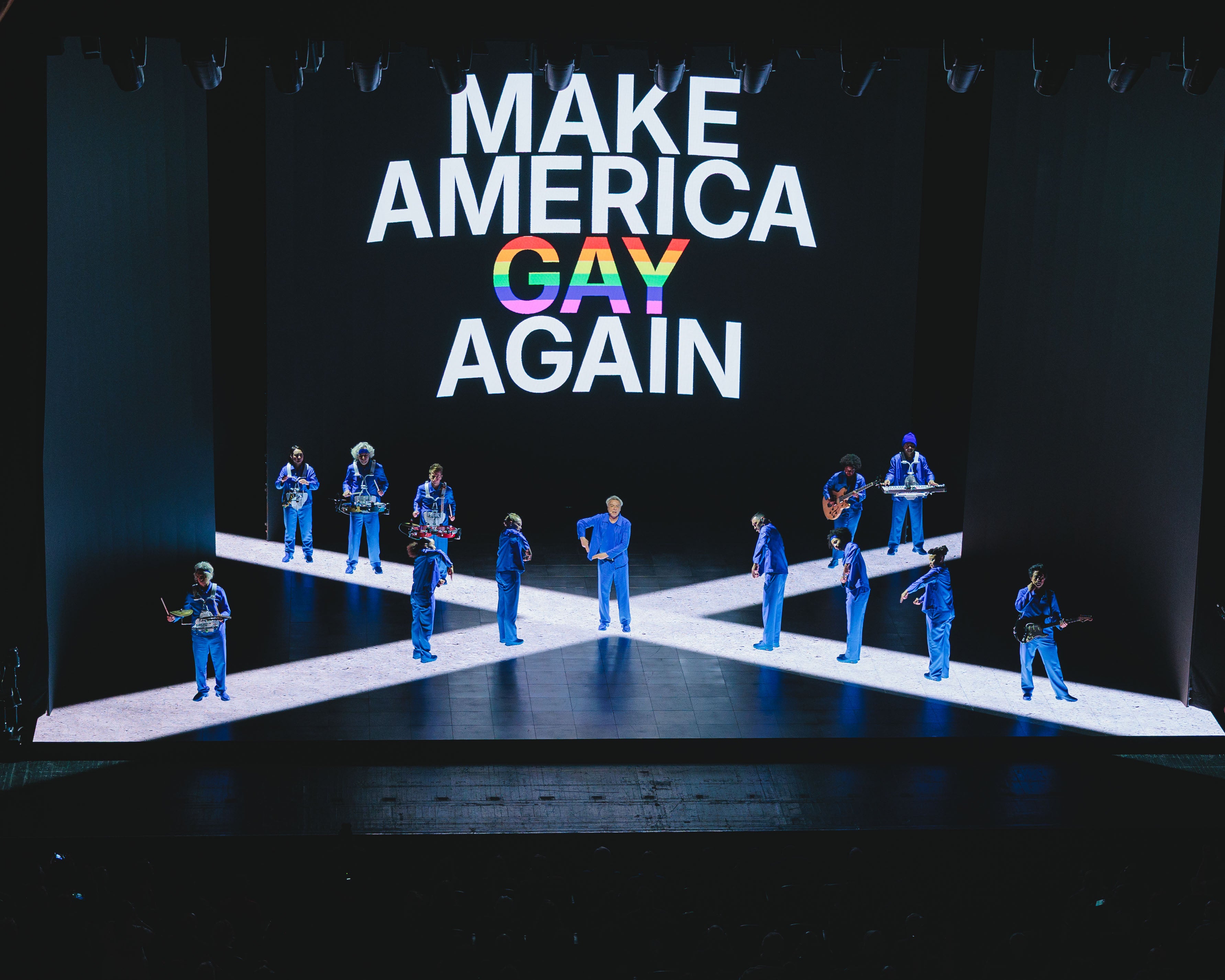 David Byrne and his band performing in front of a screen reading 'Make America Gay Again' during the song ‘T Shirt’