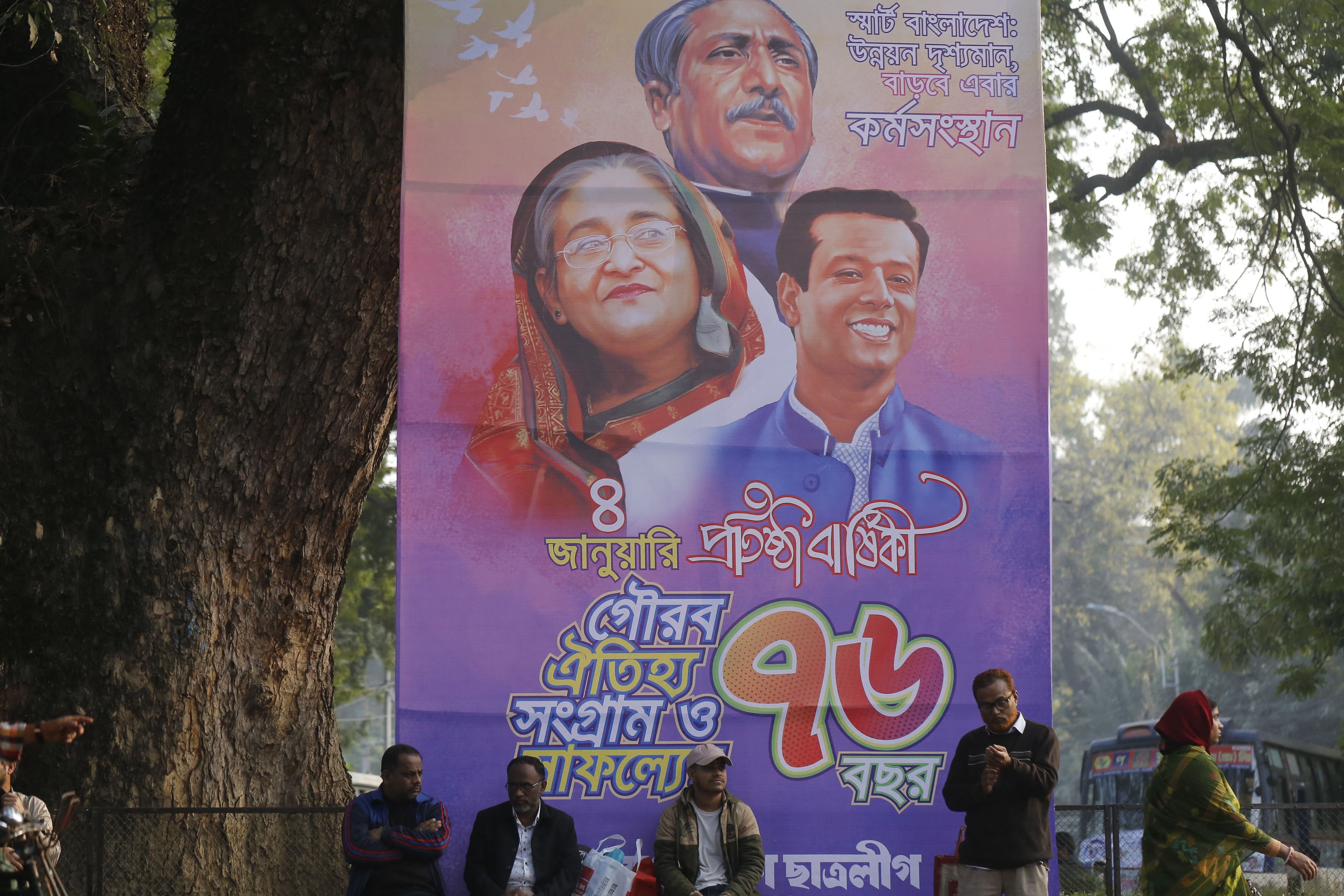 <p>People sit next to a banner marking the 74th founding anniversary of Awami League party with images of Sheikh Hasina, her father Sheikh Mujibur Rahman and son Sajeeb Wazed</p>