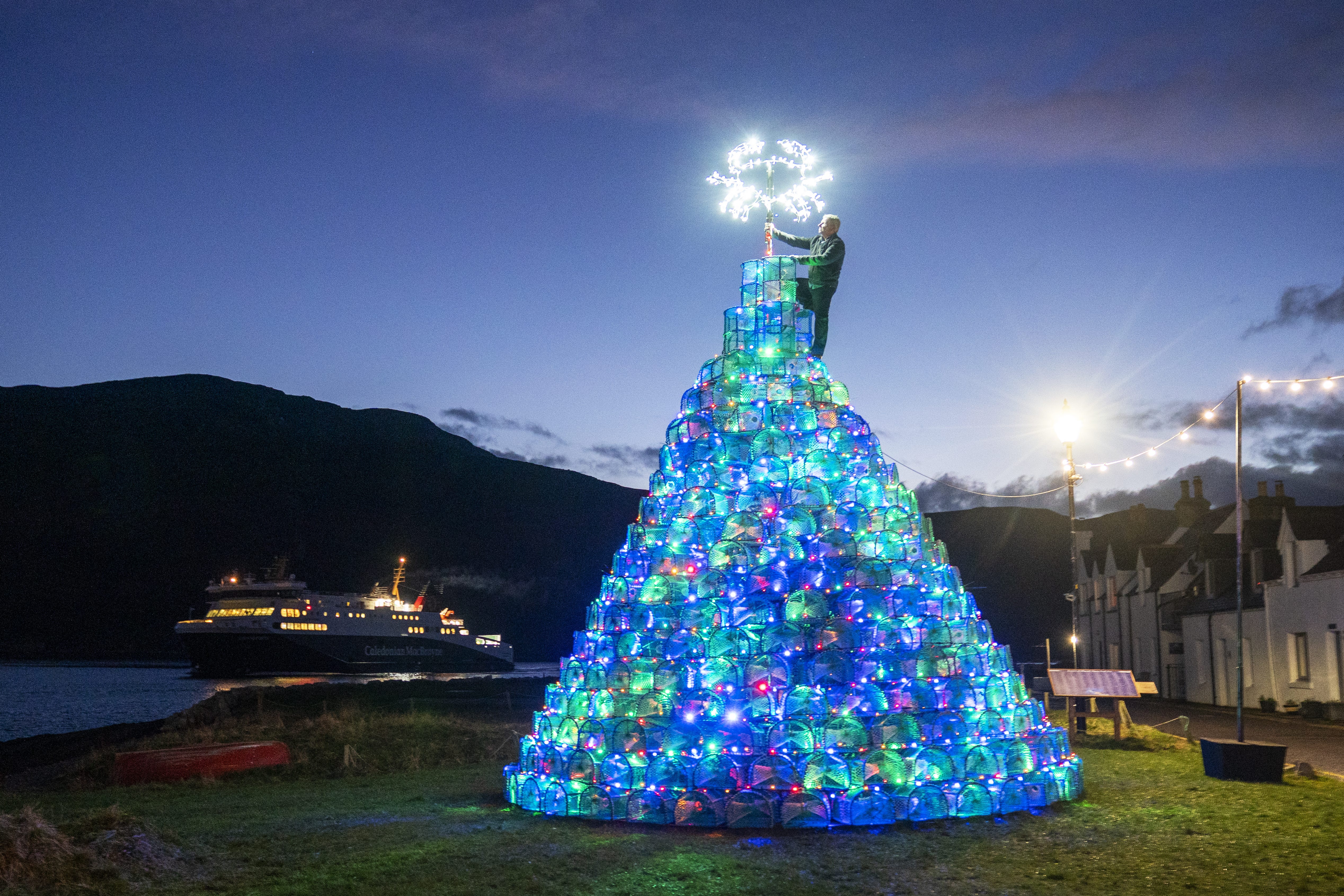 The Stornoway to Ullapool ferry sails by as Gordon Wink checks the lights on the fishing creel Christmas tree he helped to design and build on the harbour-side in Ullapool, Wester Ross. The 30ft tree, made using 340 creels used for catching crabs and prawns, has become a tradition in Ullapool since it was first constructed in 2016 (Jane Barlow/PA)